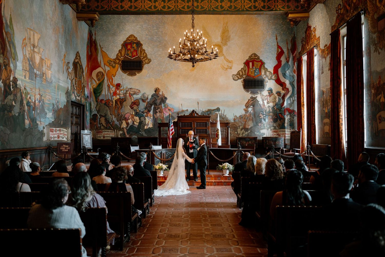Wide shot of elegant wedding ceremony in historic hall with colorful murals and seated guests.