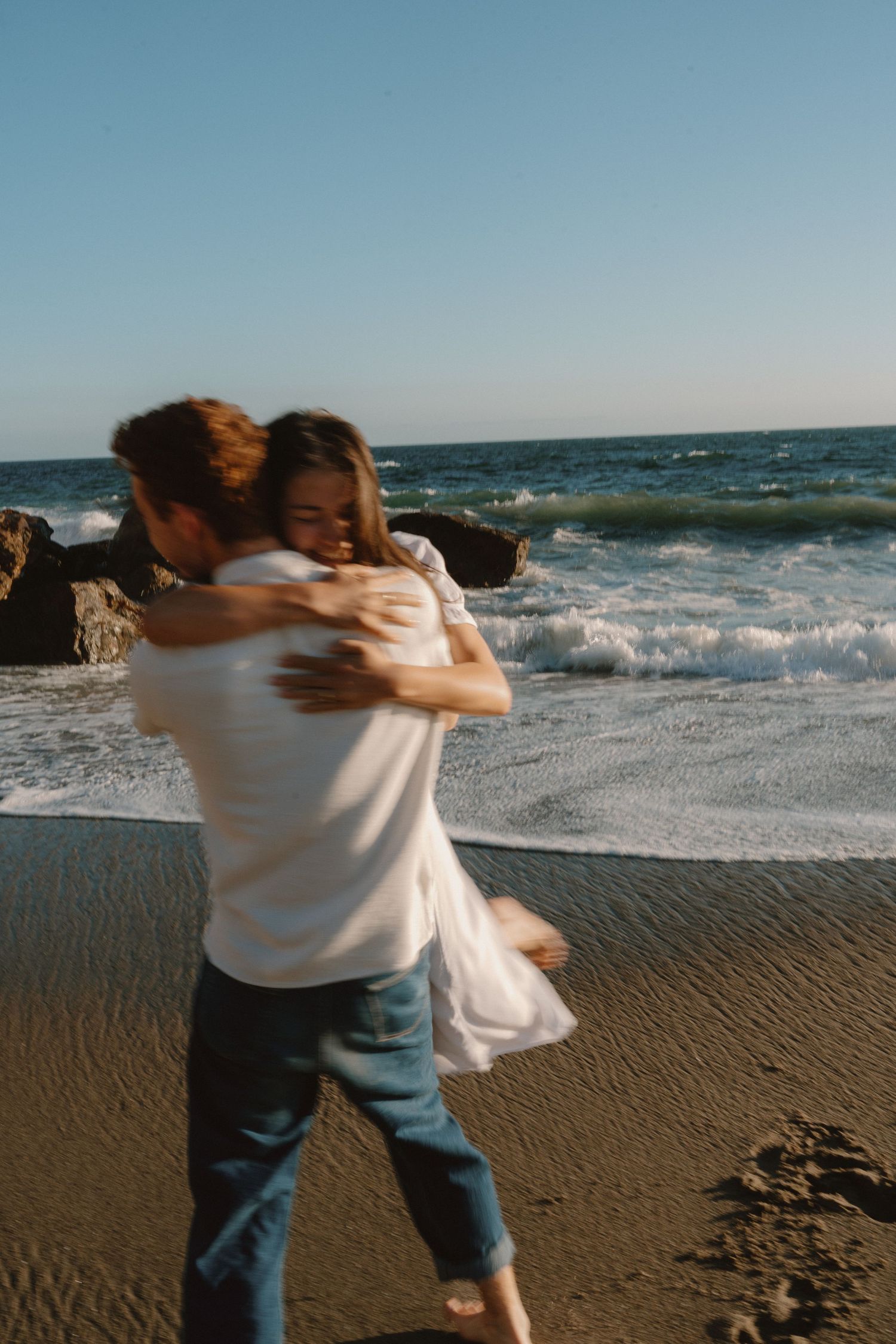 Couple embracing and spinning on a sandy beach with ocean waves and rocks in the background.