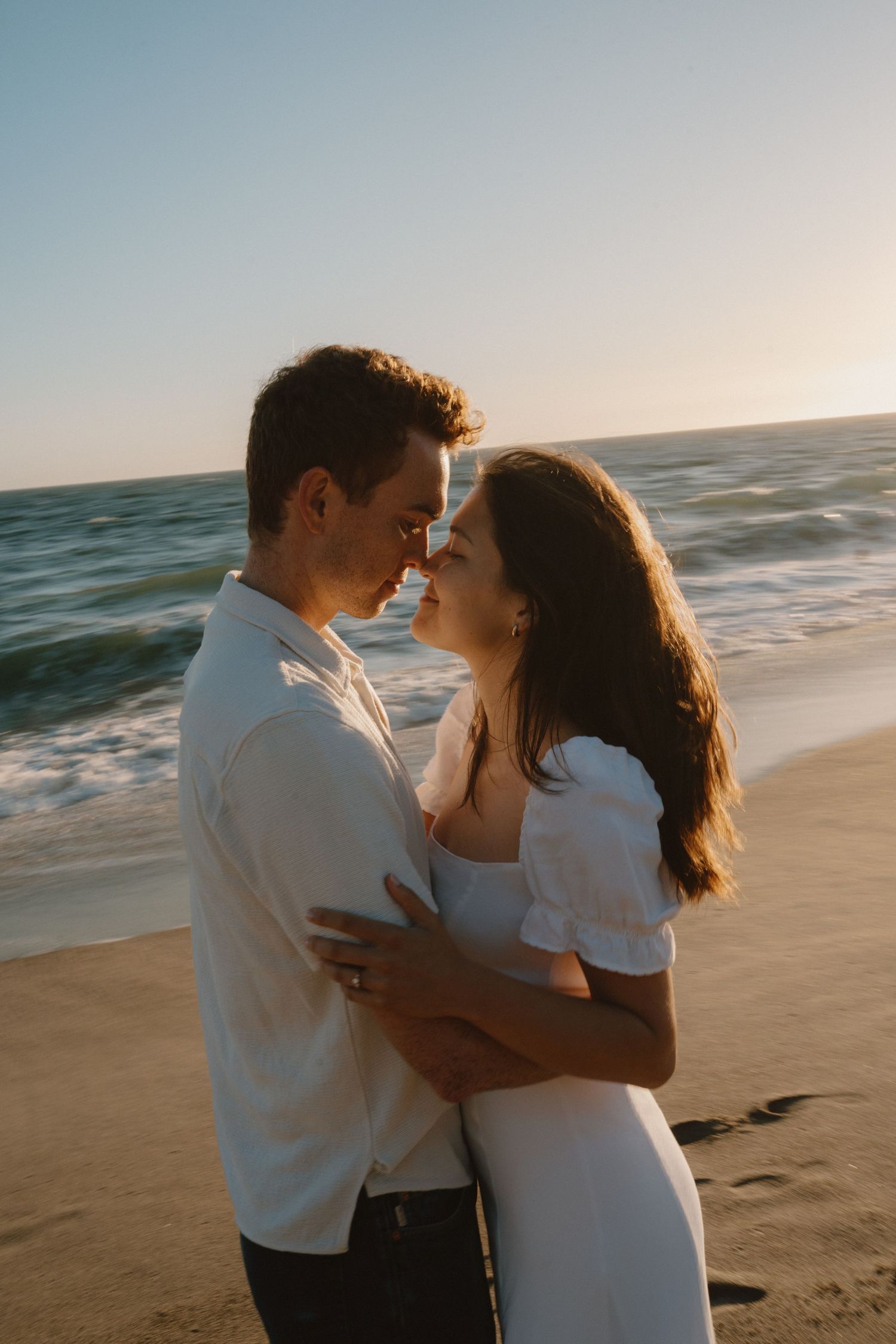 Couple sharing a tender almost-kiss embrace on a beach at golden hour with ocean waves behind them.