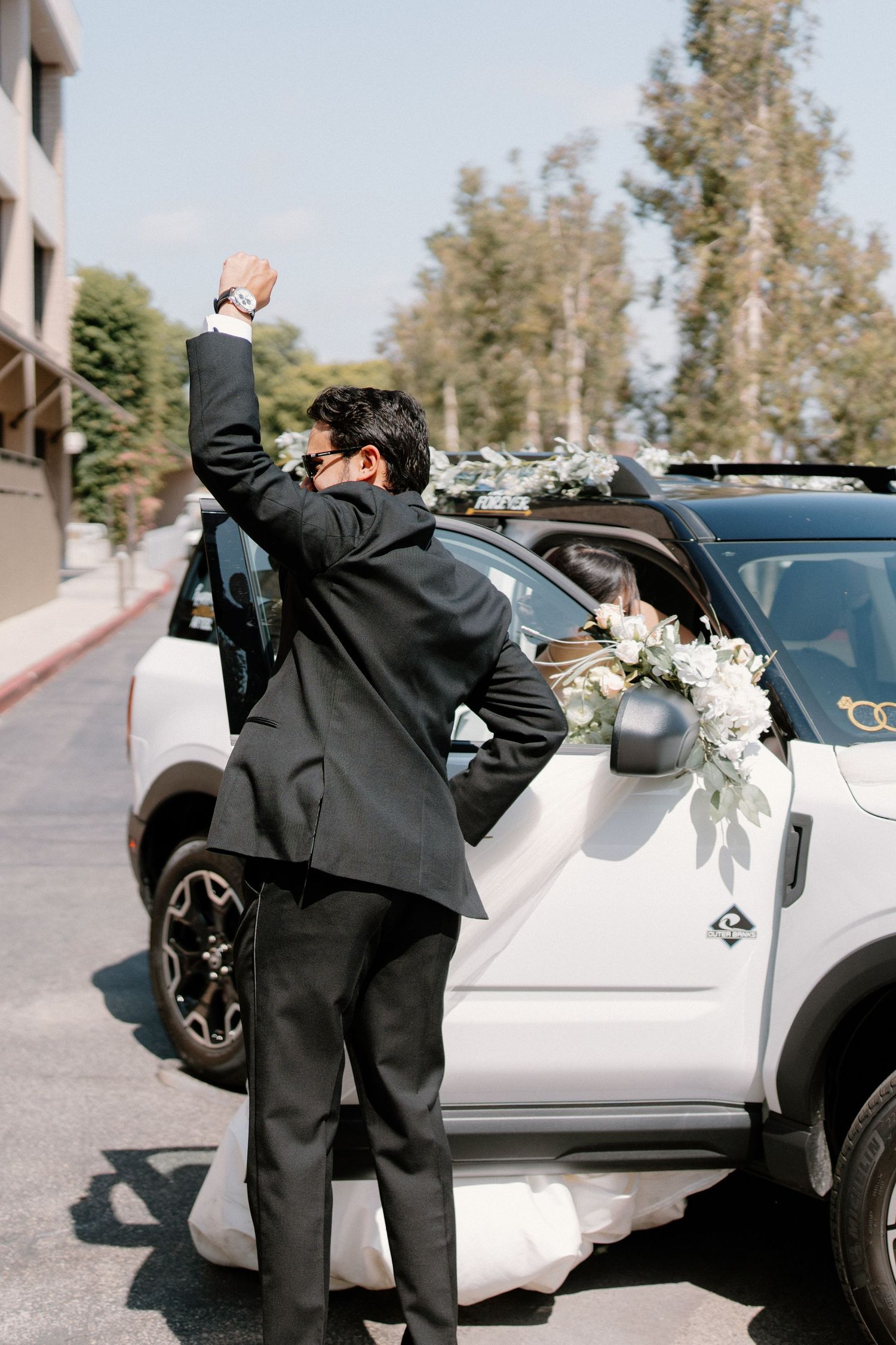 Groom in black suit raising fist in celebration beside a white SUV decorated with floral greenery.