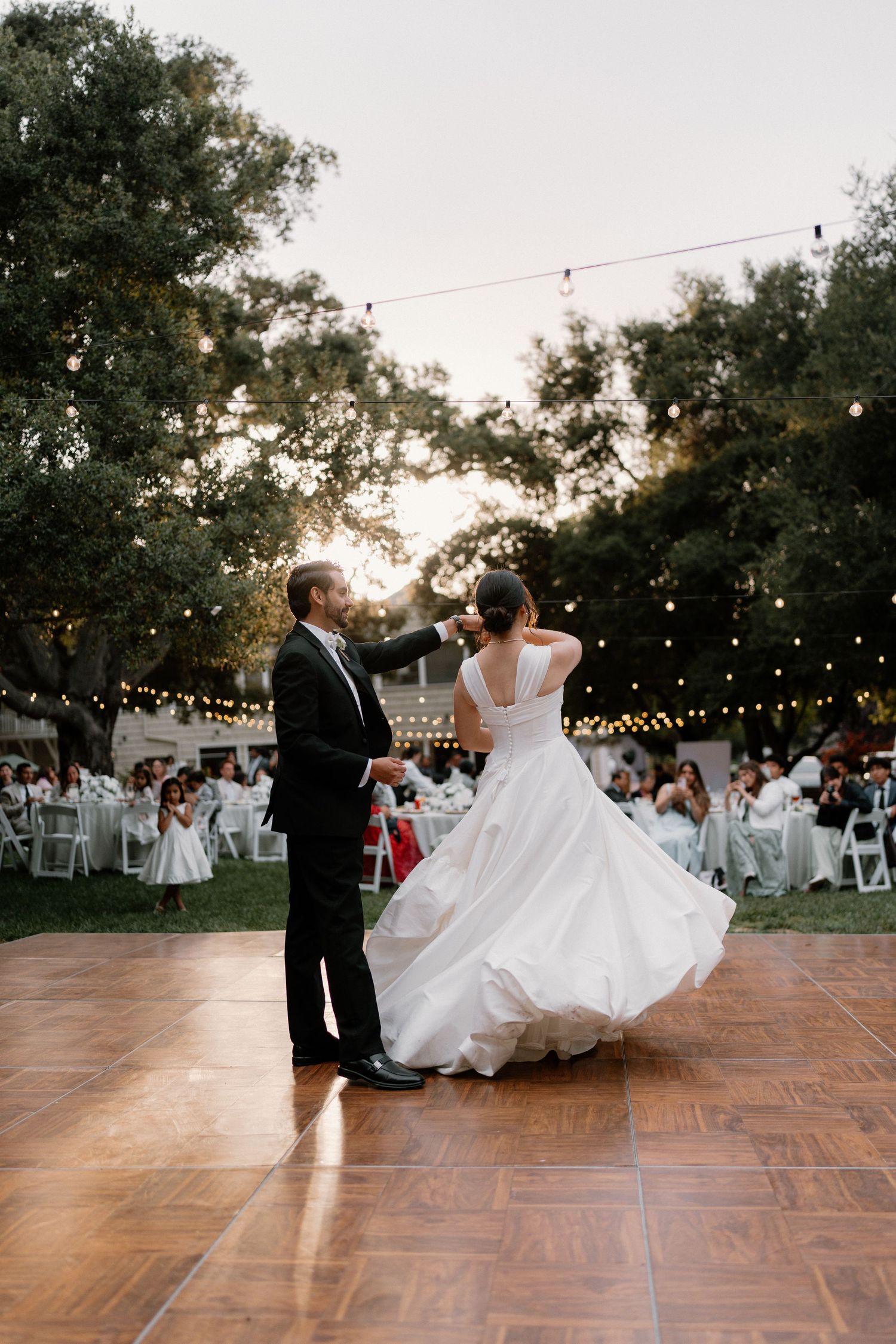 Bride twirls in flowing white ball gown as groom leads their first dance under string lights at sunset.
