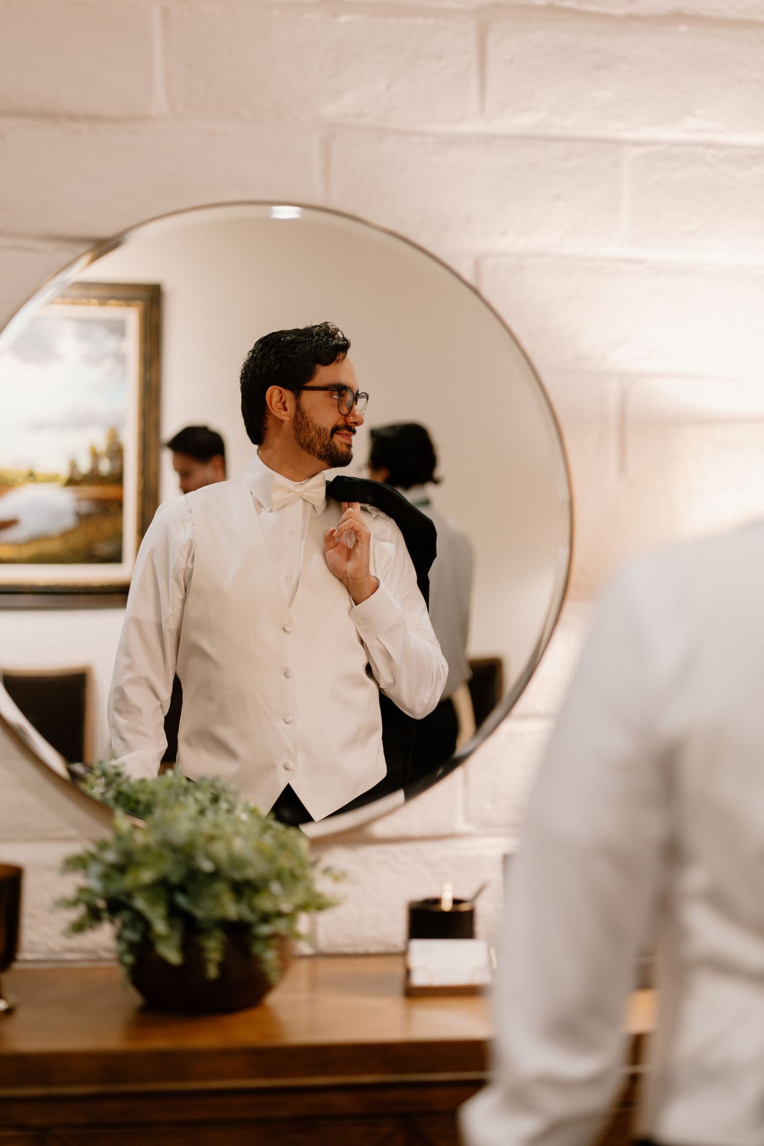 Groom in white vest adjusting tie while looking in round mirror during wedding day preparations.