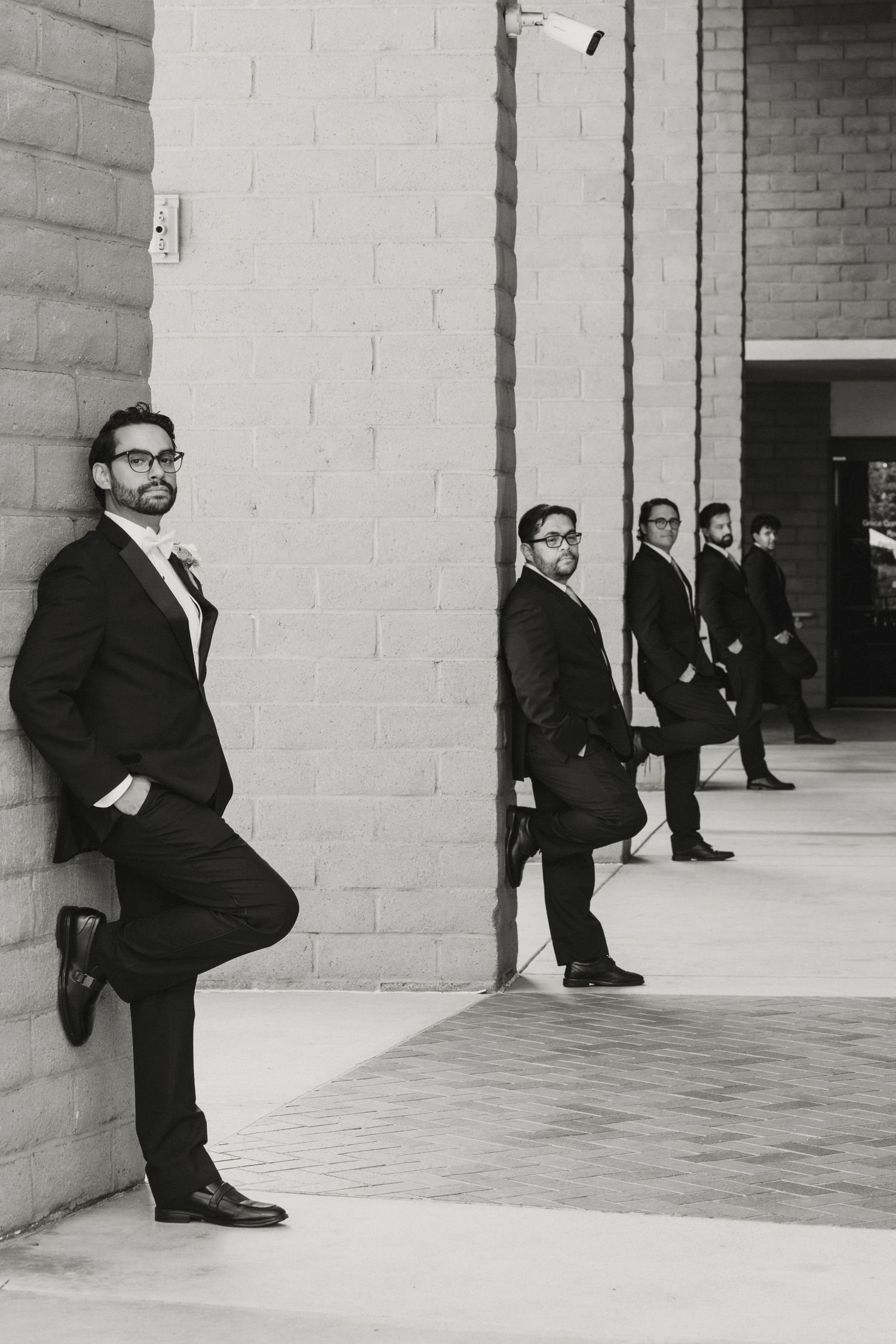 Black and white photo of groomsmen in suits leaning against a brick wall outside a building.