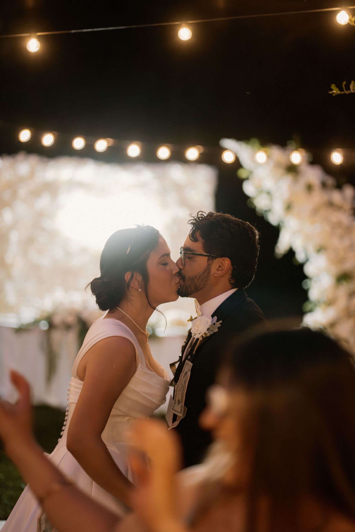 Bride and groom share their first kiss at an outdoor evening wedding ceremony under string lights.