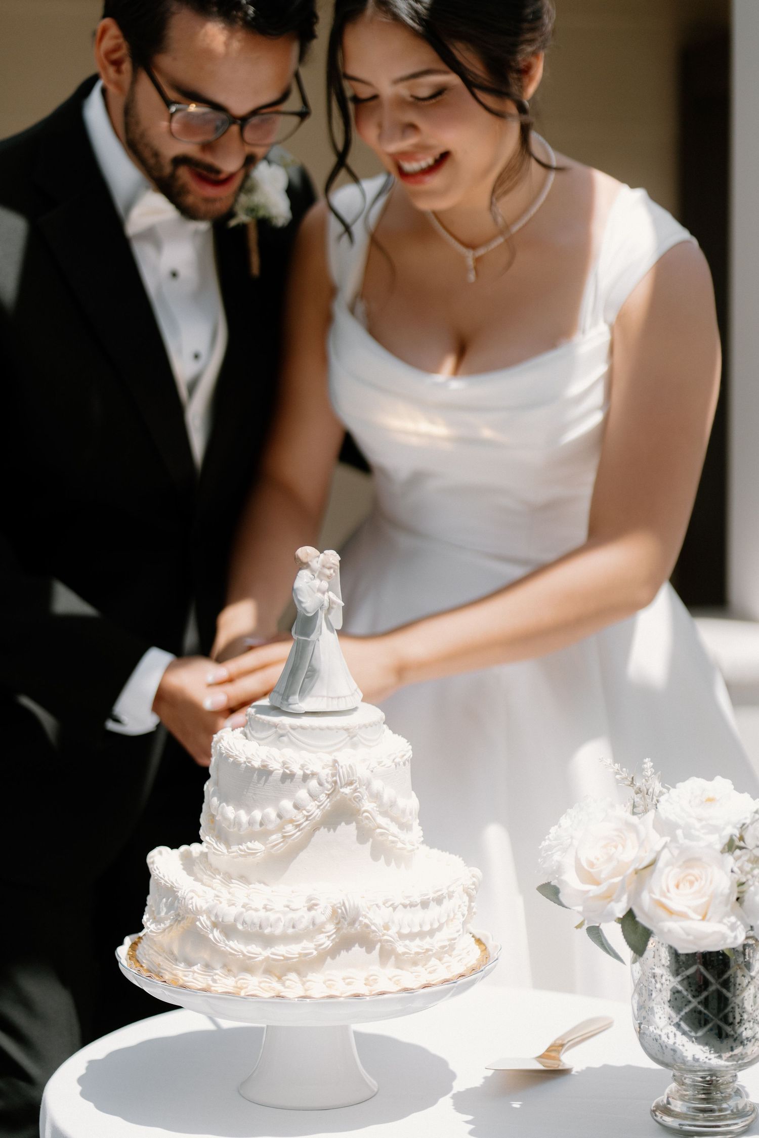 Bride and groom cut elegant two-tier white wedding cake topped with classic bride and groom figurines.