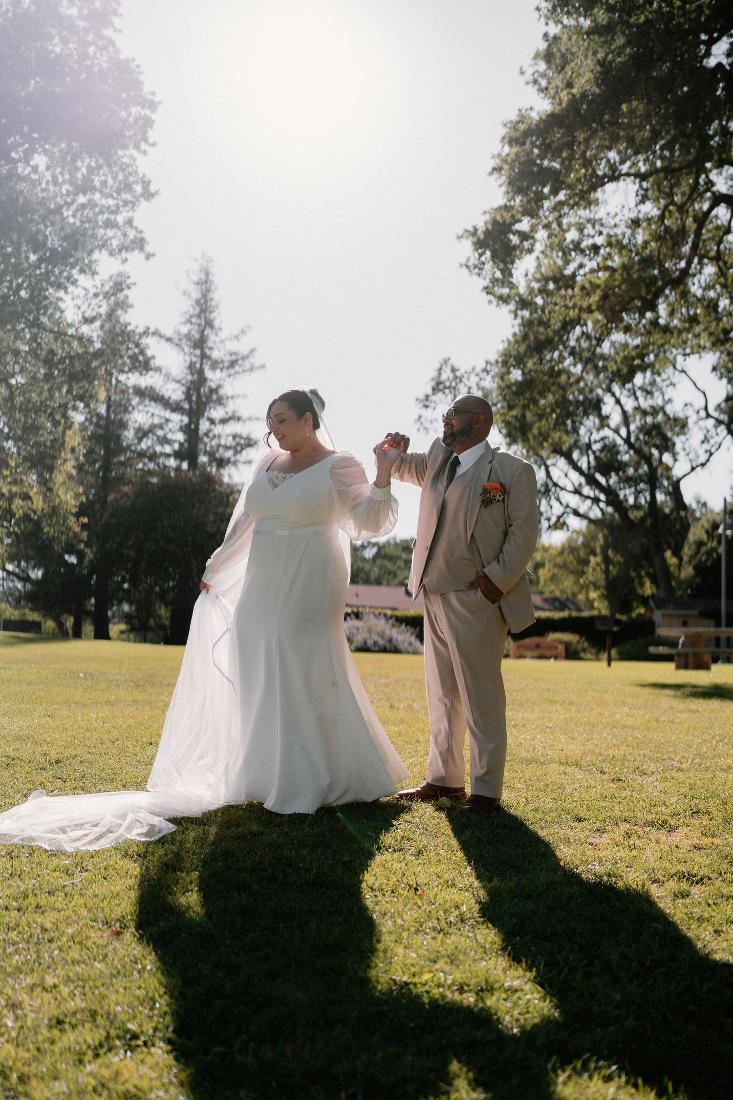 Bride in white gown and groom in beige suit dance joyfully on a sunny green lawn.