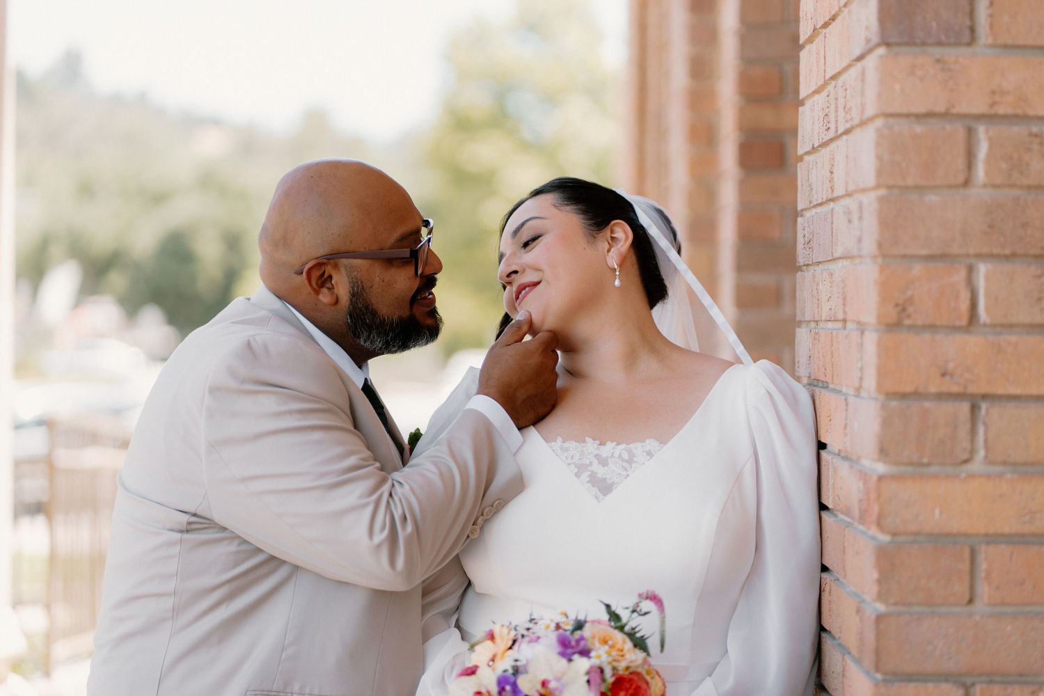 Groom in tan suit tenderly touches brides chin as she smiles holding colorful bouquet by brick wall.