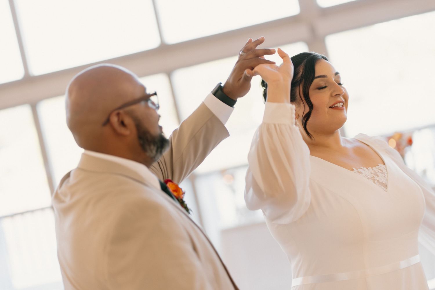 Groom in tan suit spinning bride in white dress during joyful first dance at wedding reception.