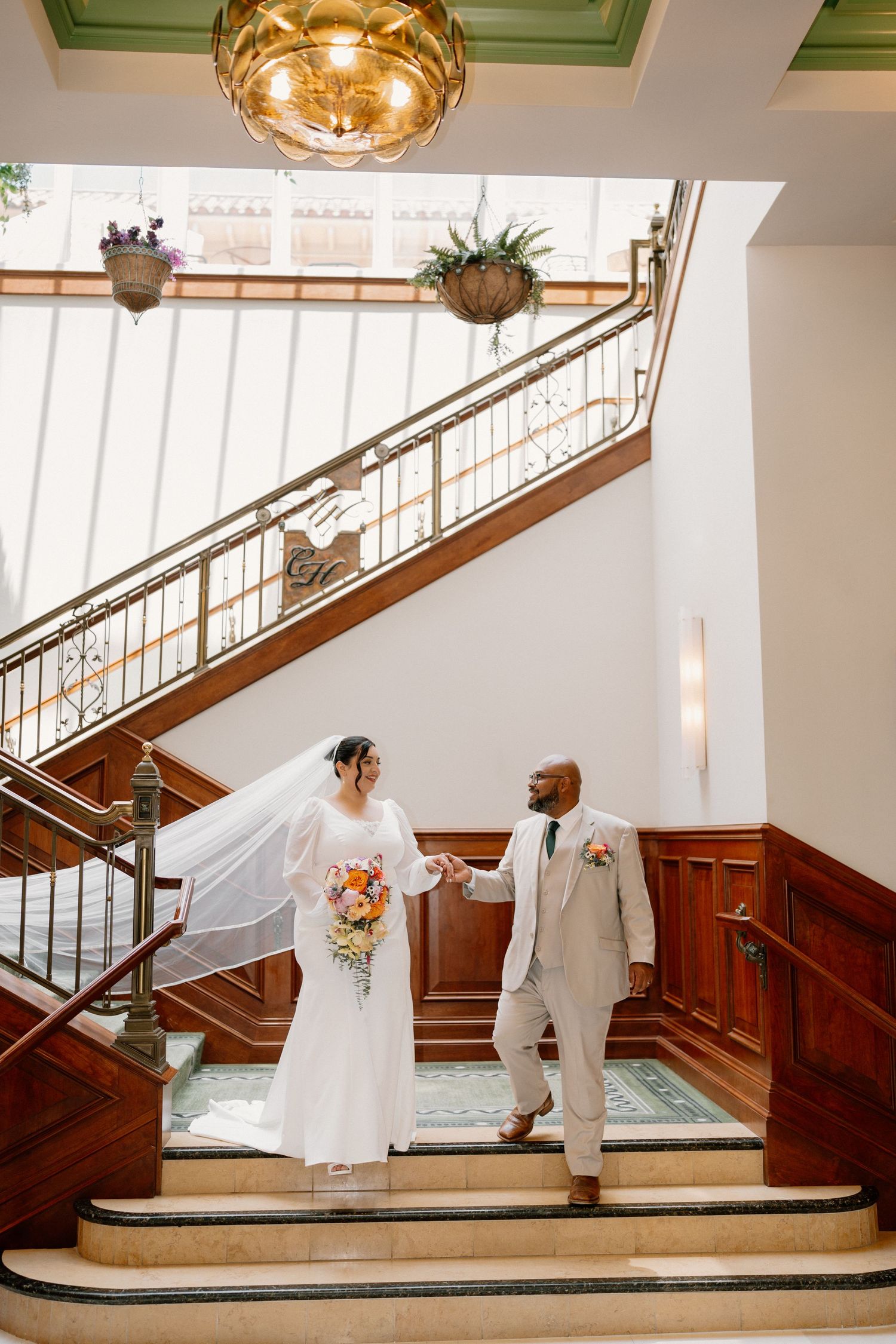 Bride and groom hold hands on elegant staircase, bride in white gown with veil holding bouquet, groom in tan suit.