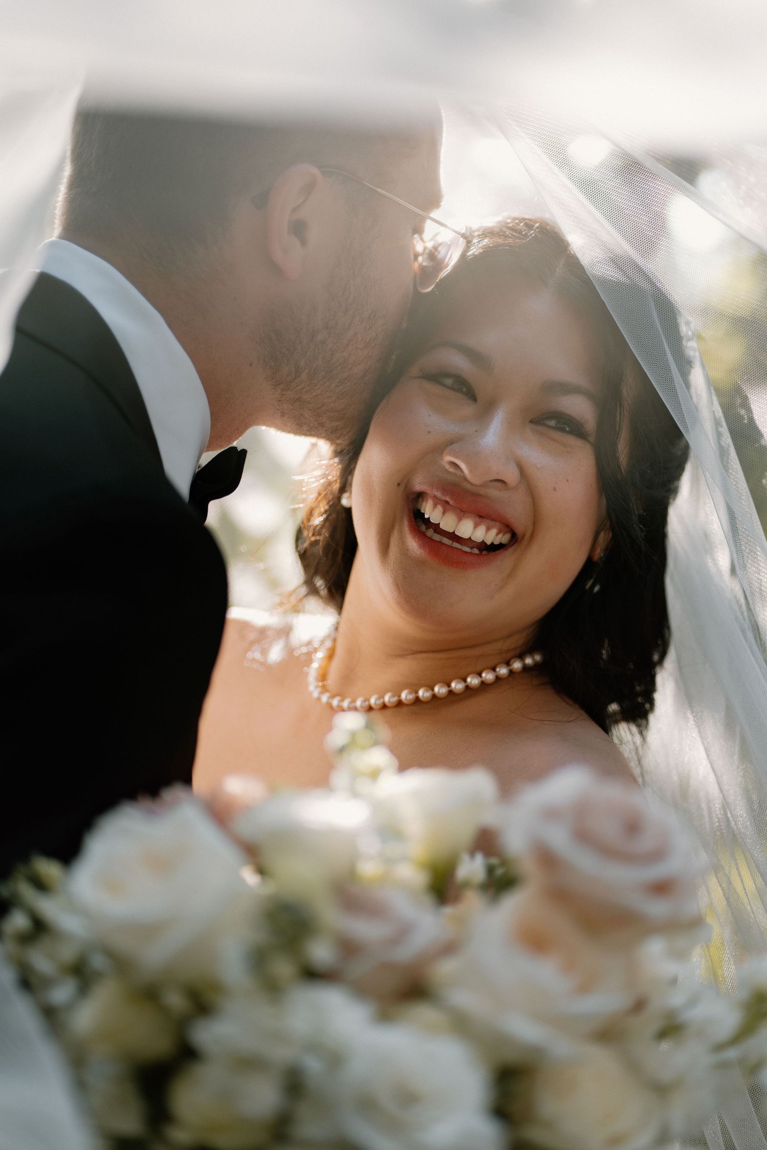 Joyful bride laughing under veil with groom, holding white rose bouquet on wedding day.