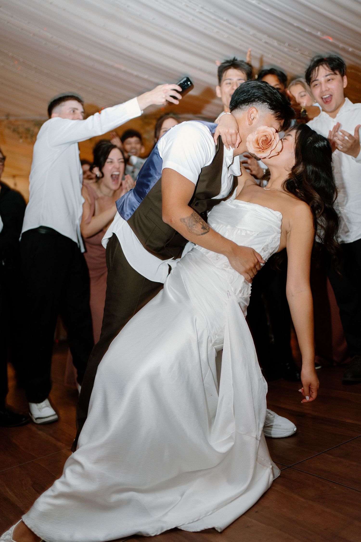 Groom dips bride for passionate kiss on dance floor surrounded by cheering guests at wedding reception.