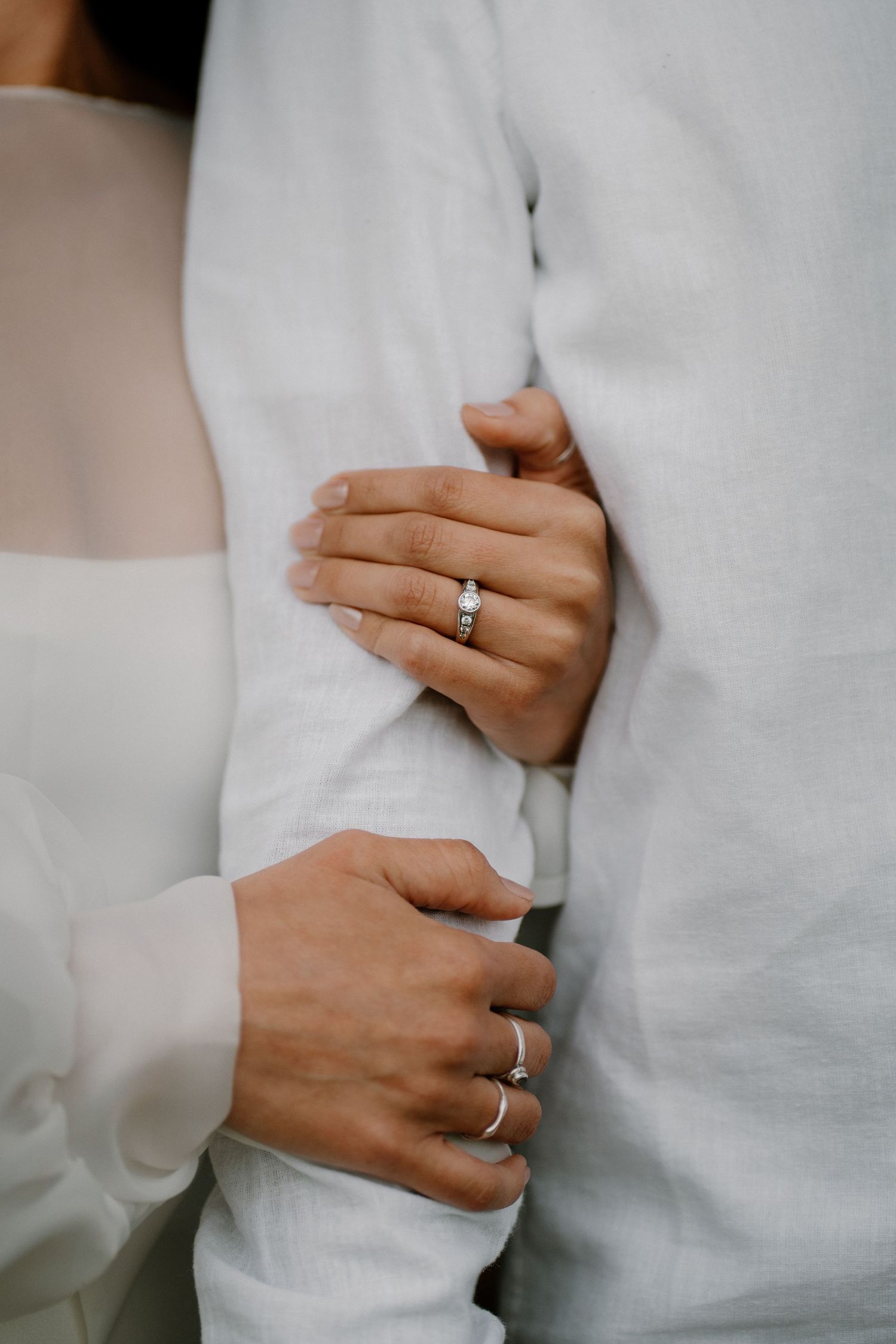 Close-up of a womans hand with a diamond engagement ring resting on a mans white linen shirt.