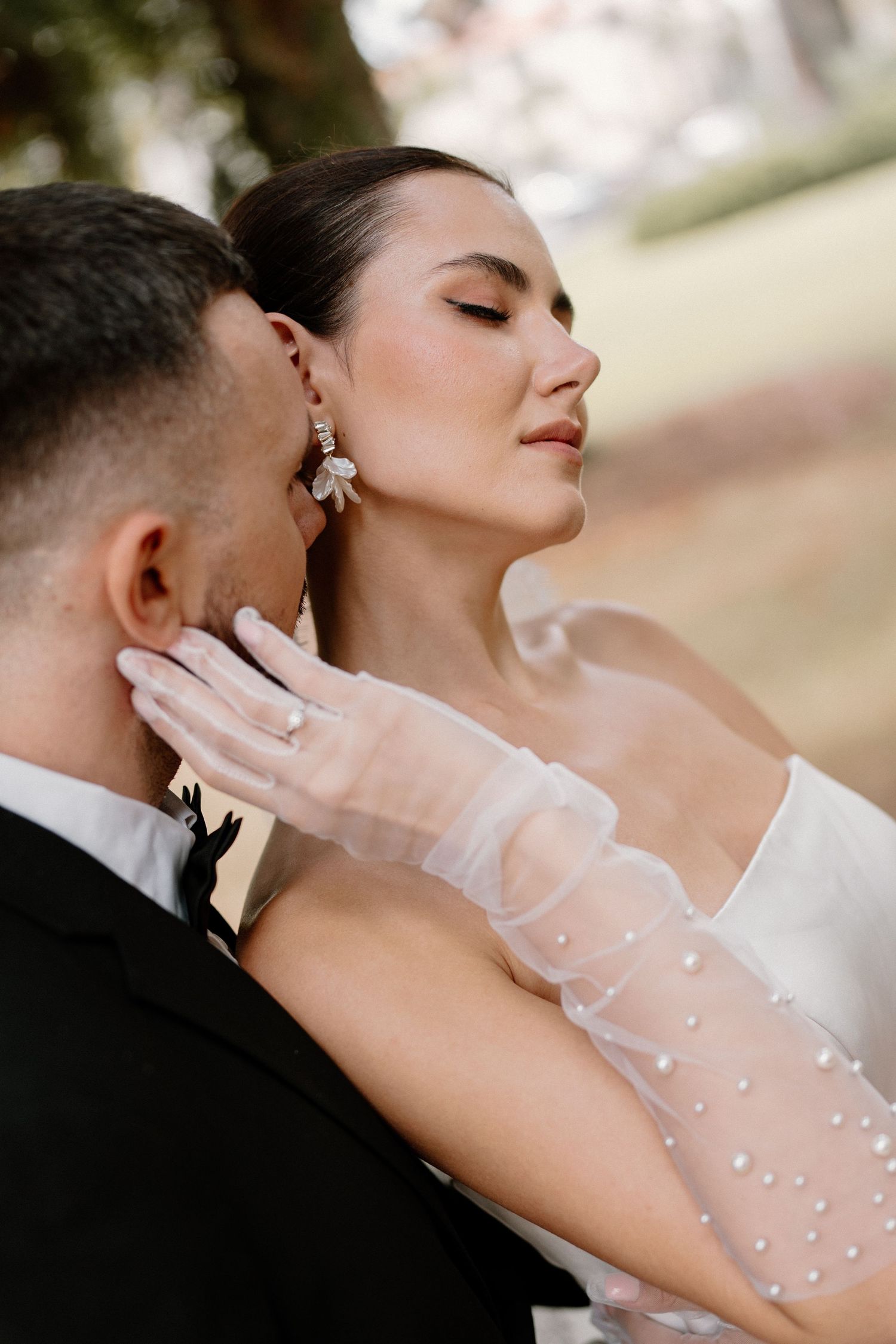 Bride in off-shoulder gown with pearl gloves tenderly embracing groom outdoors.