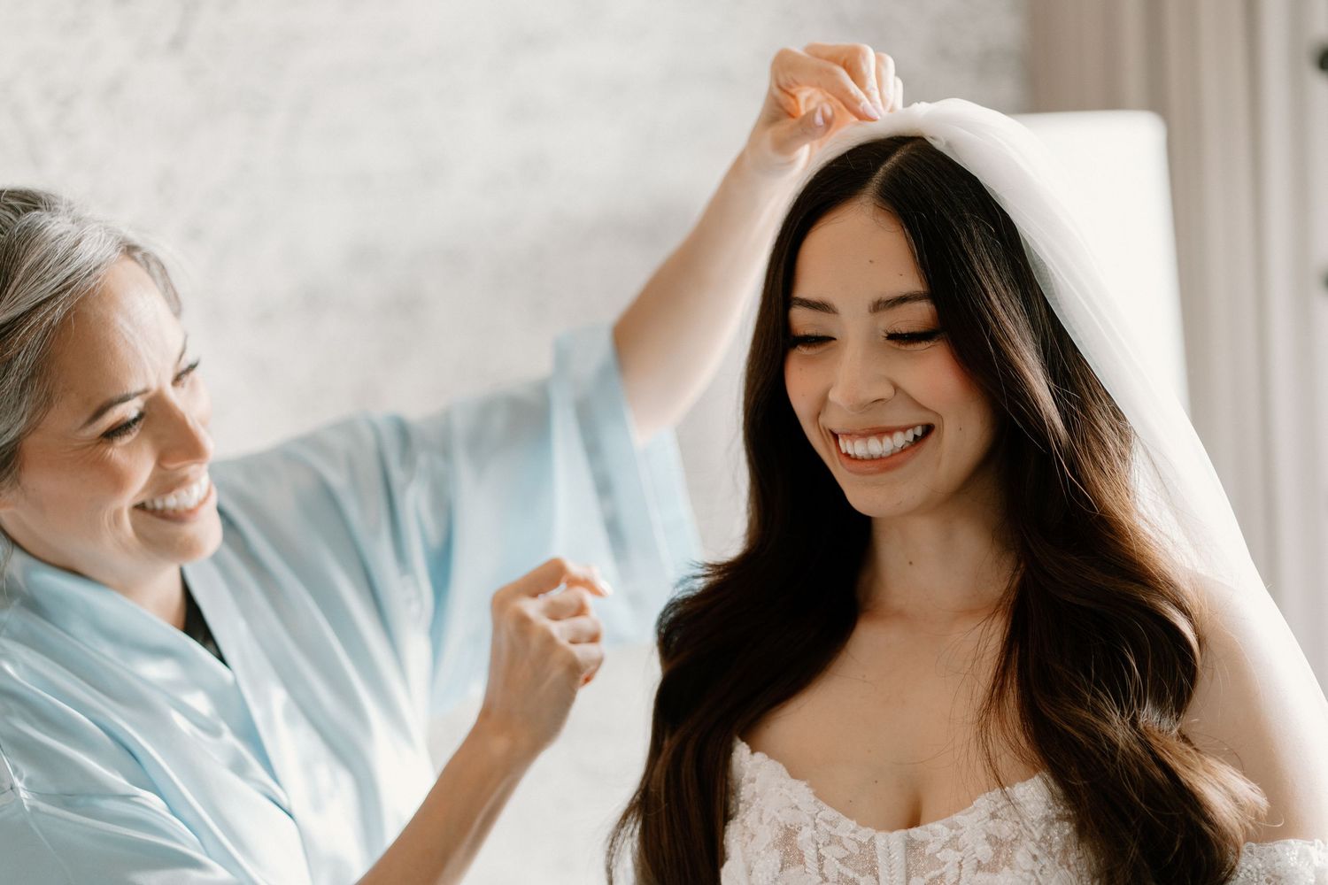 Smiling bride having her wedding veil placed by a woman in a light blue robe during bridal preparations.