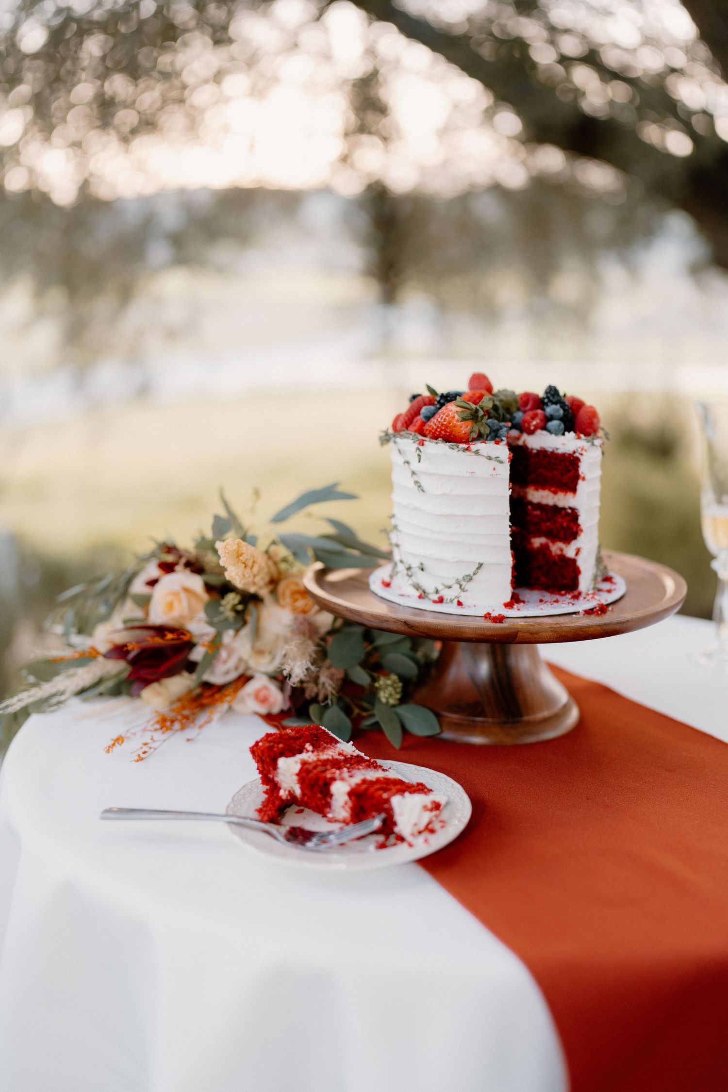 Sliced red velvet wedding cake with white frosting and fresh berries on wooden stand with bouquet.