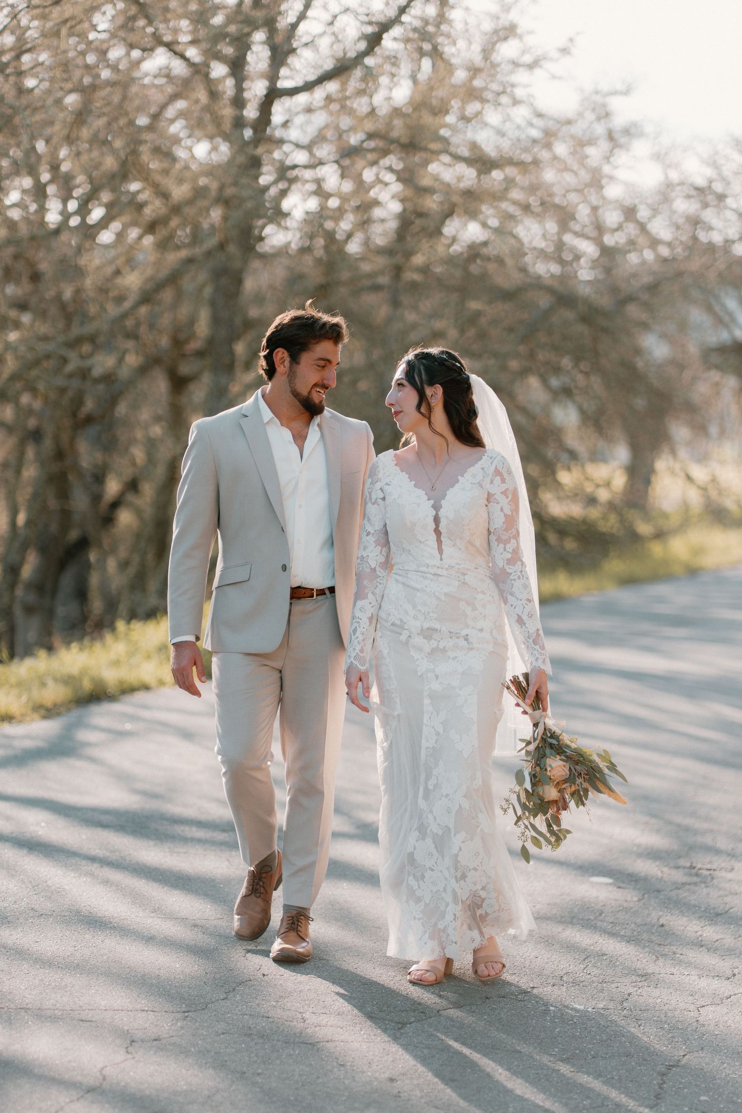 A smiling bride and groom walk hand in hand down a sunlit road on their wedding day.