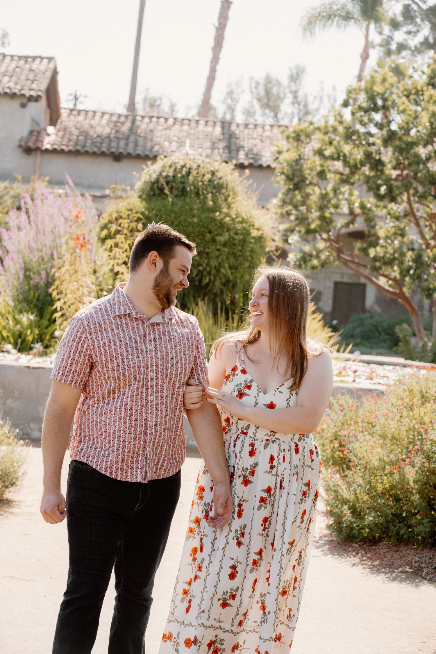 A smiling couple walks hand-in-hand through a sunny garden with colorful flowers and a Spanish-style building.