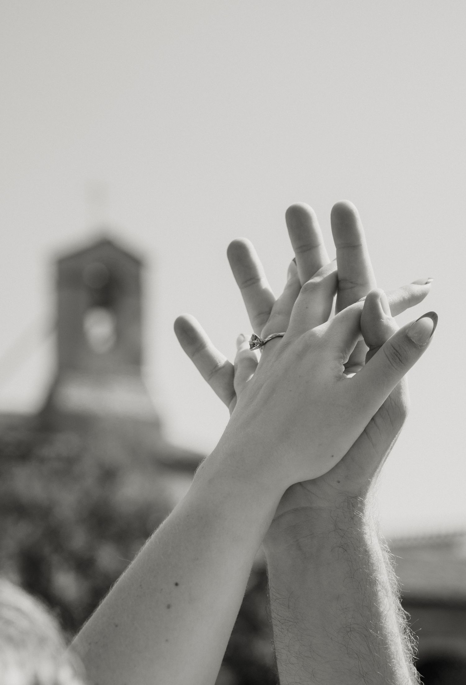 Black and white photo of two hands raised together, wearing rings, with a church tower in the background.