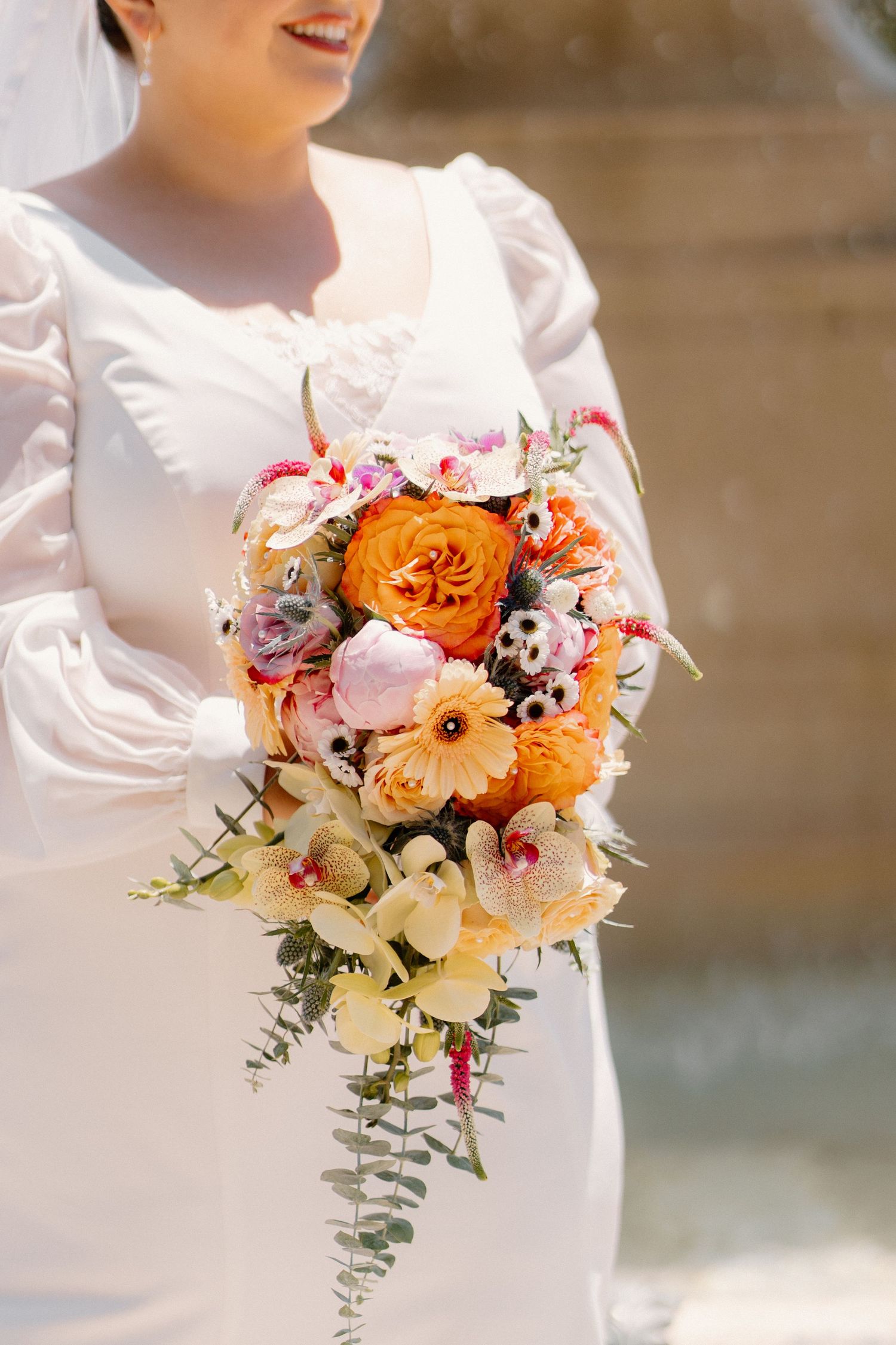 Bride in white dress holding vibrant orange, pink, and cream cascading wedding bouquet with eucalyptus.