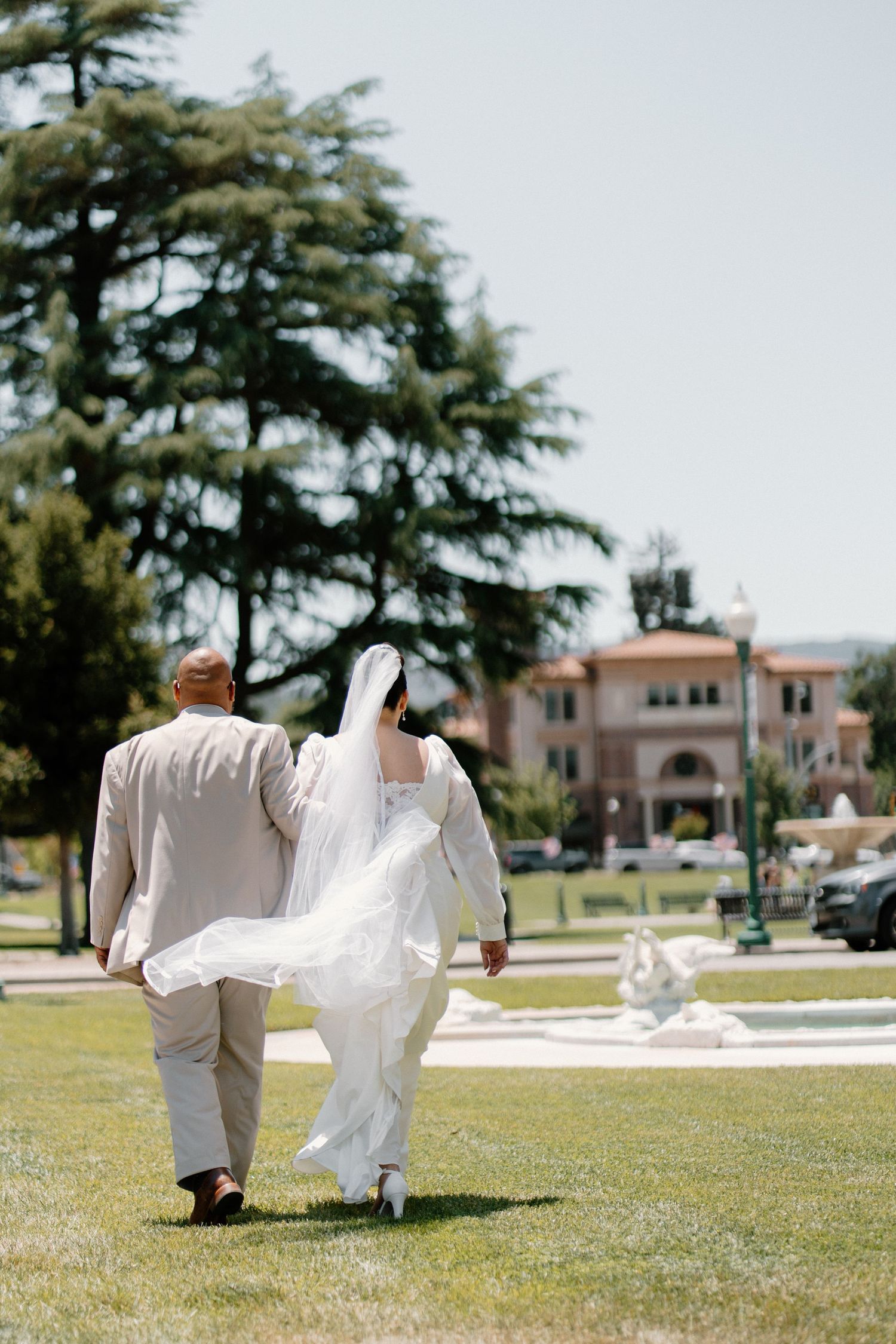 Bride and groom walk hand-in-arm across green lawn with large trees and elegant venue building in background.