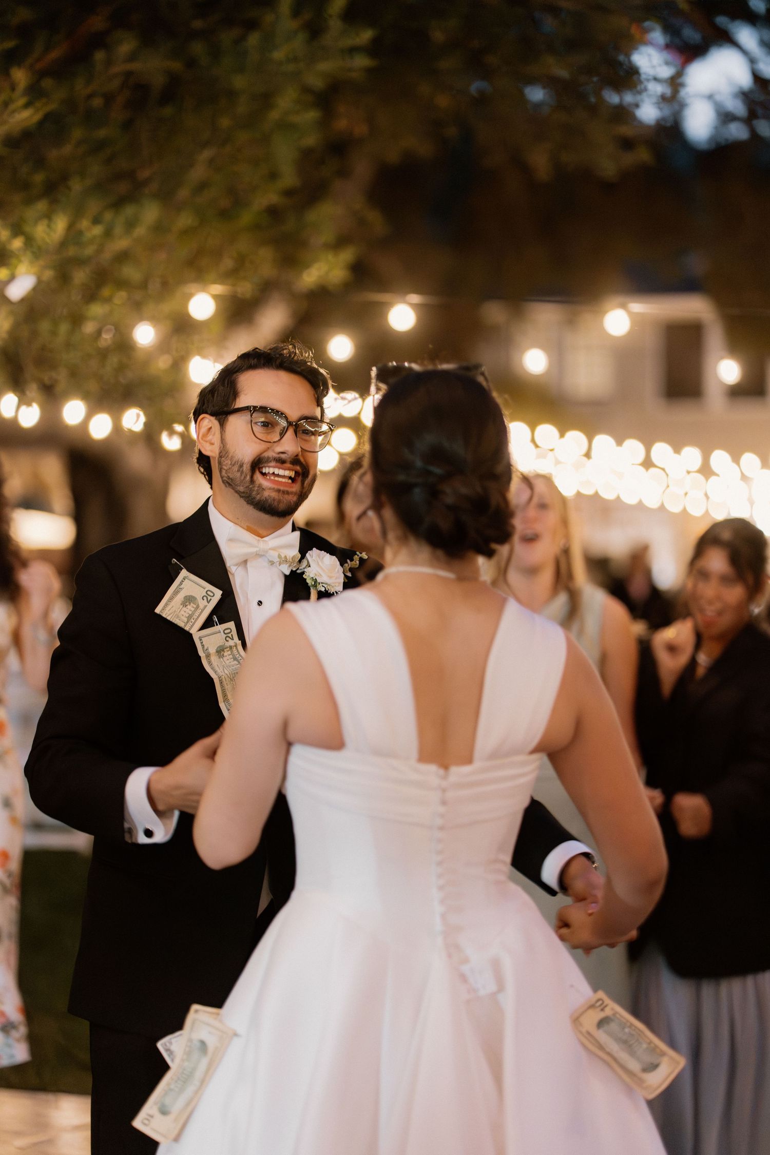 Smiling groom in tuxedo dances with bride under string lights, with money tucked in his jacket.