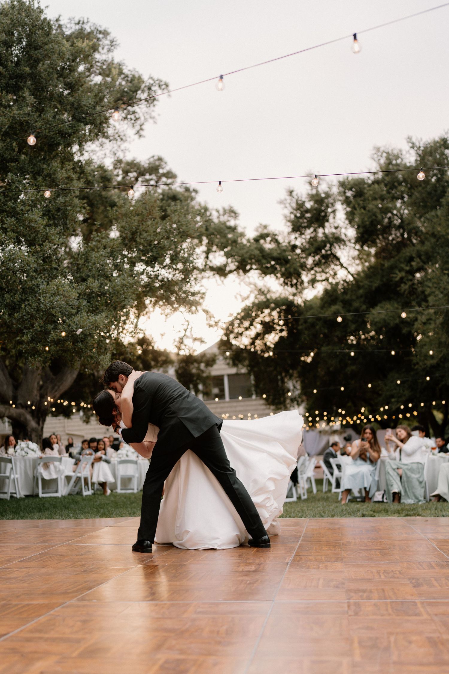 Groom dips bride dramatically on outdoor dance floor under string lights at twilight reception.