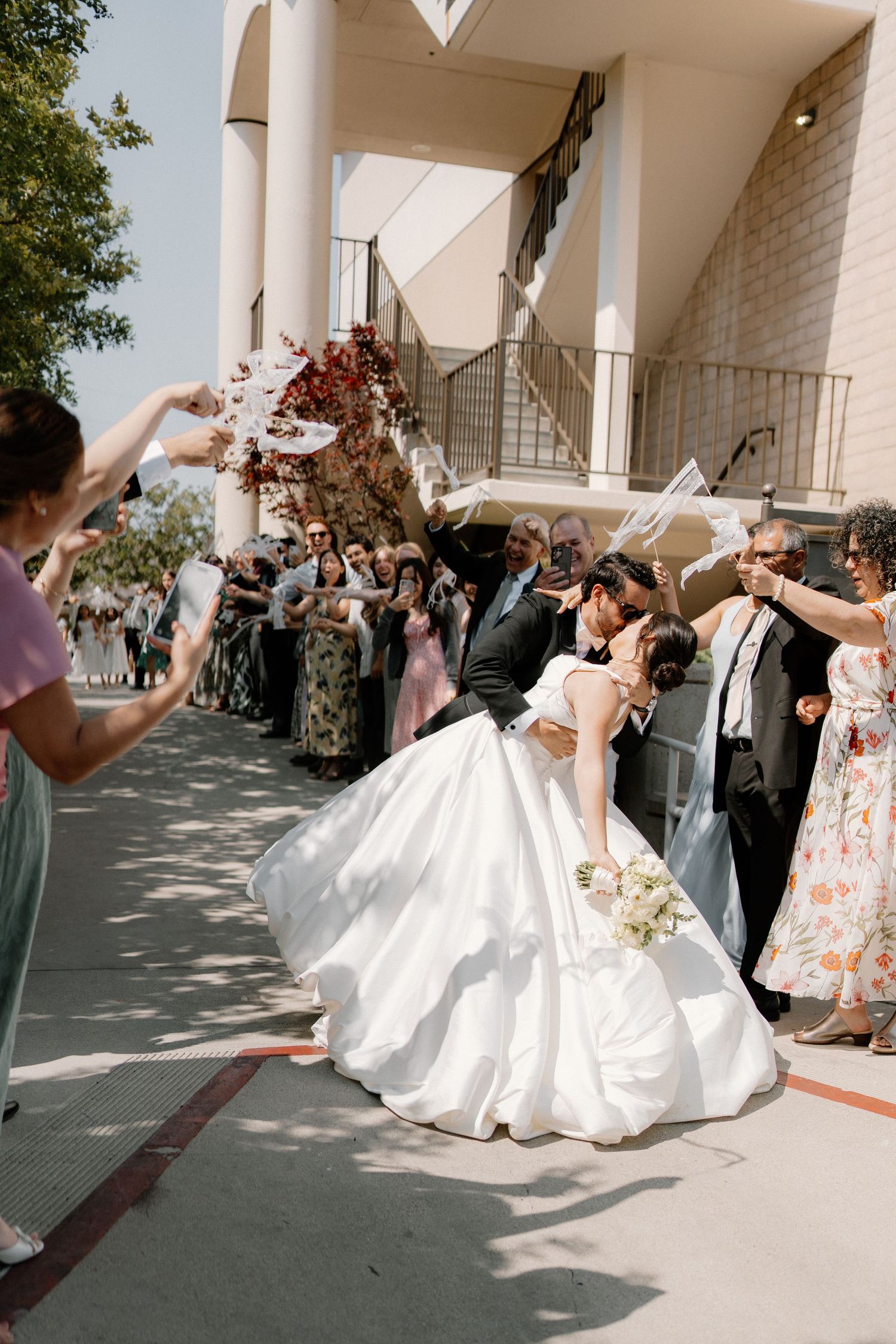 Groom dips bride for a kiss outside church entrance as wedding guests celebrate and cheer around them.