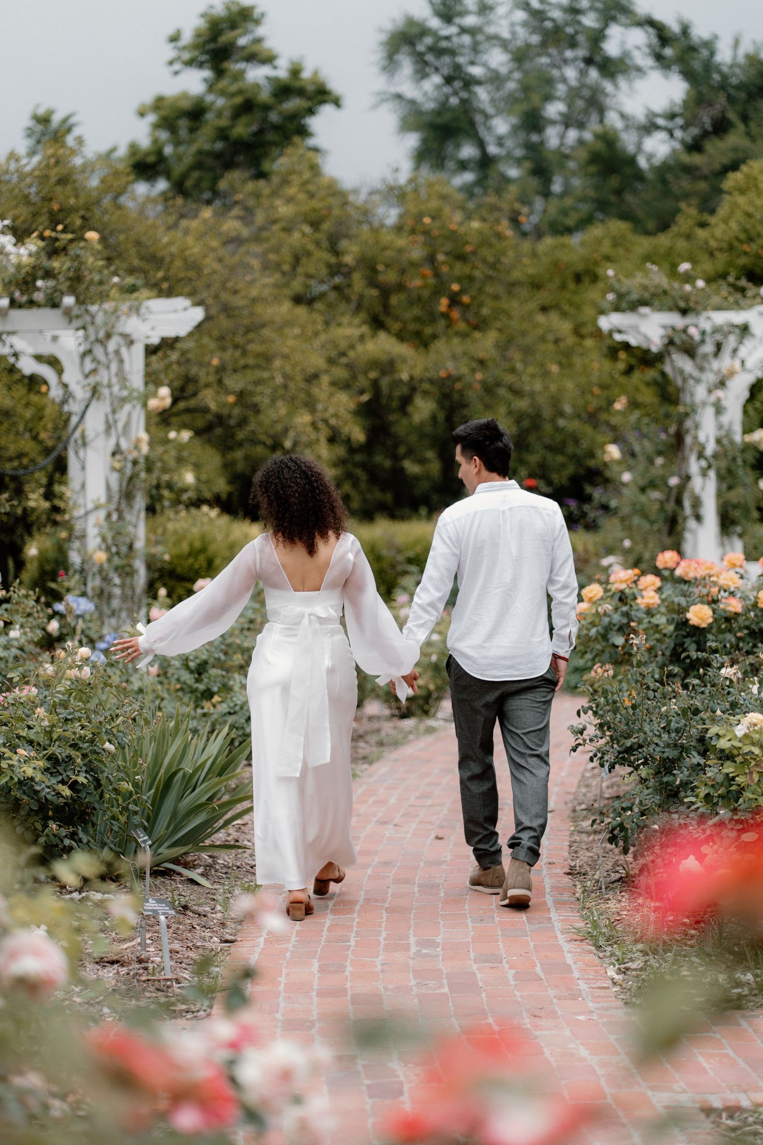 Couple holding hands walking down a brick garden path surrounded by colorful roses and white arbors.