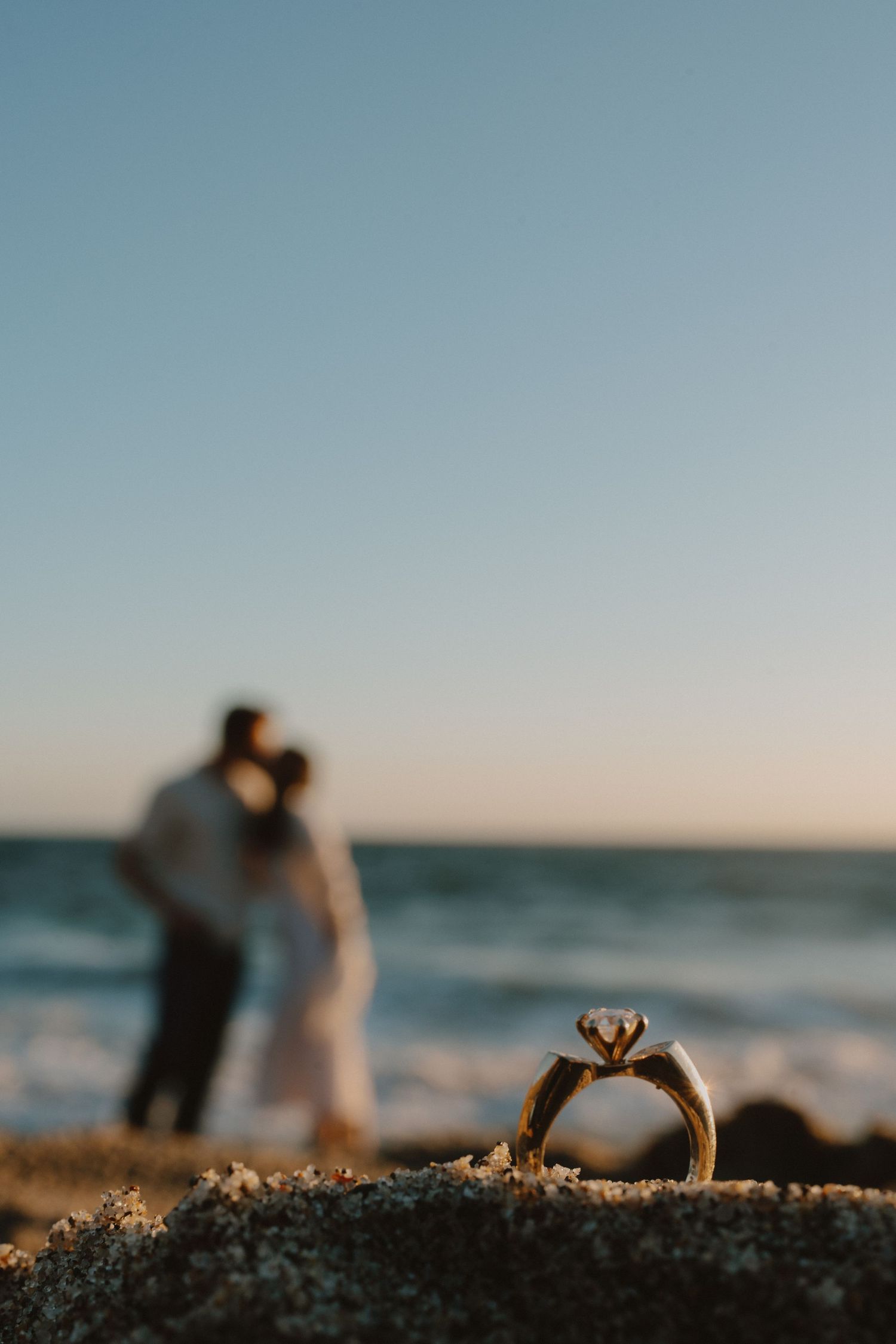 Diamond engagement ring in focus on sandy beach with couple kissing in blurred background at sunset.