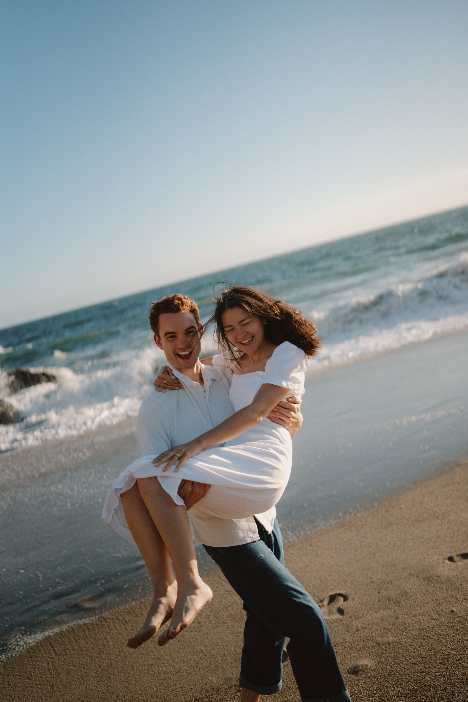 A joyful couple laughing on a beach, man carrying woman in his arms near ocean waves.