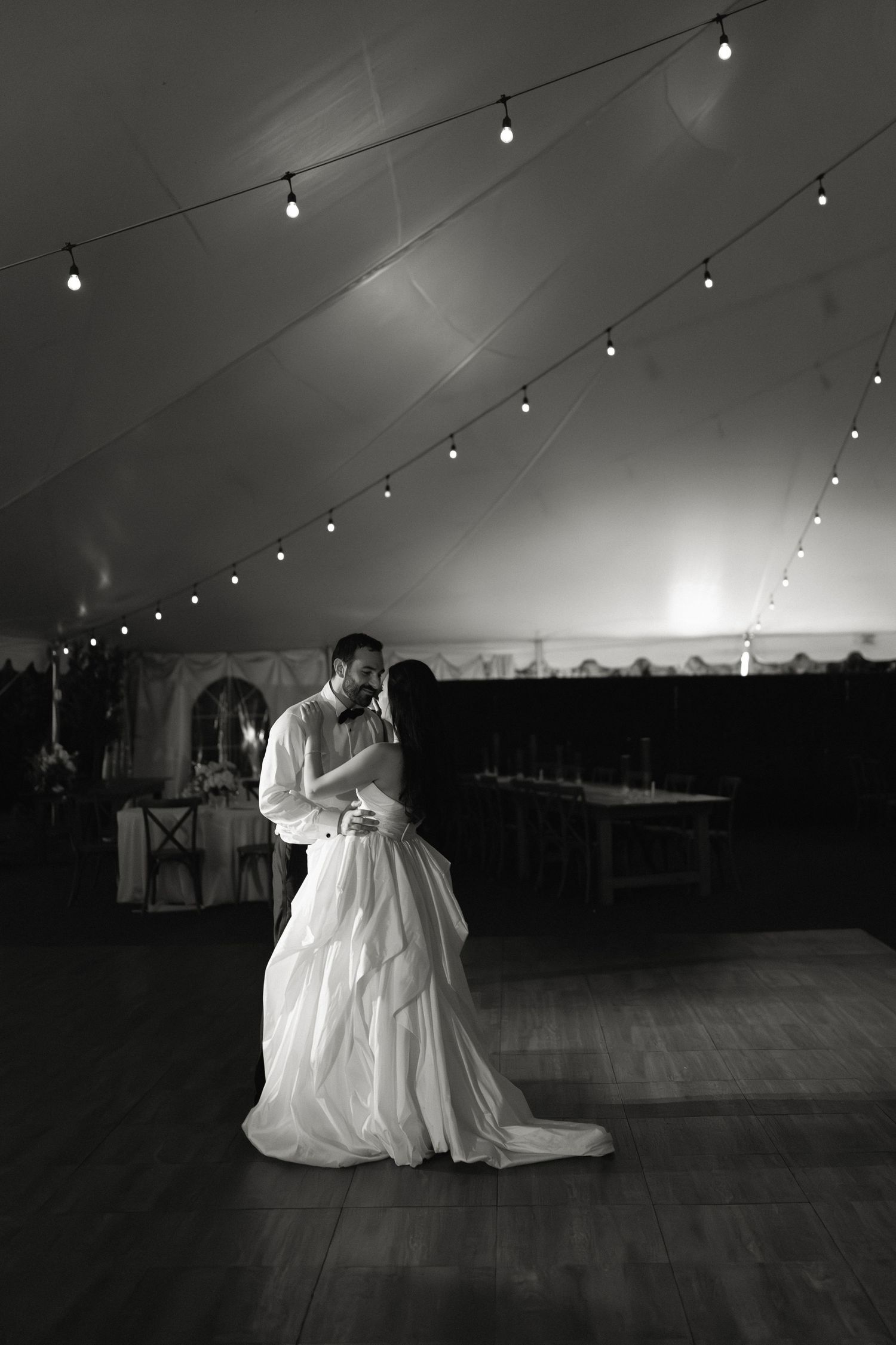 Black and white photo of bride and groom sharing first dance under string lights in a tent.