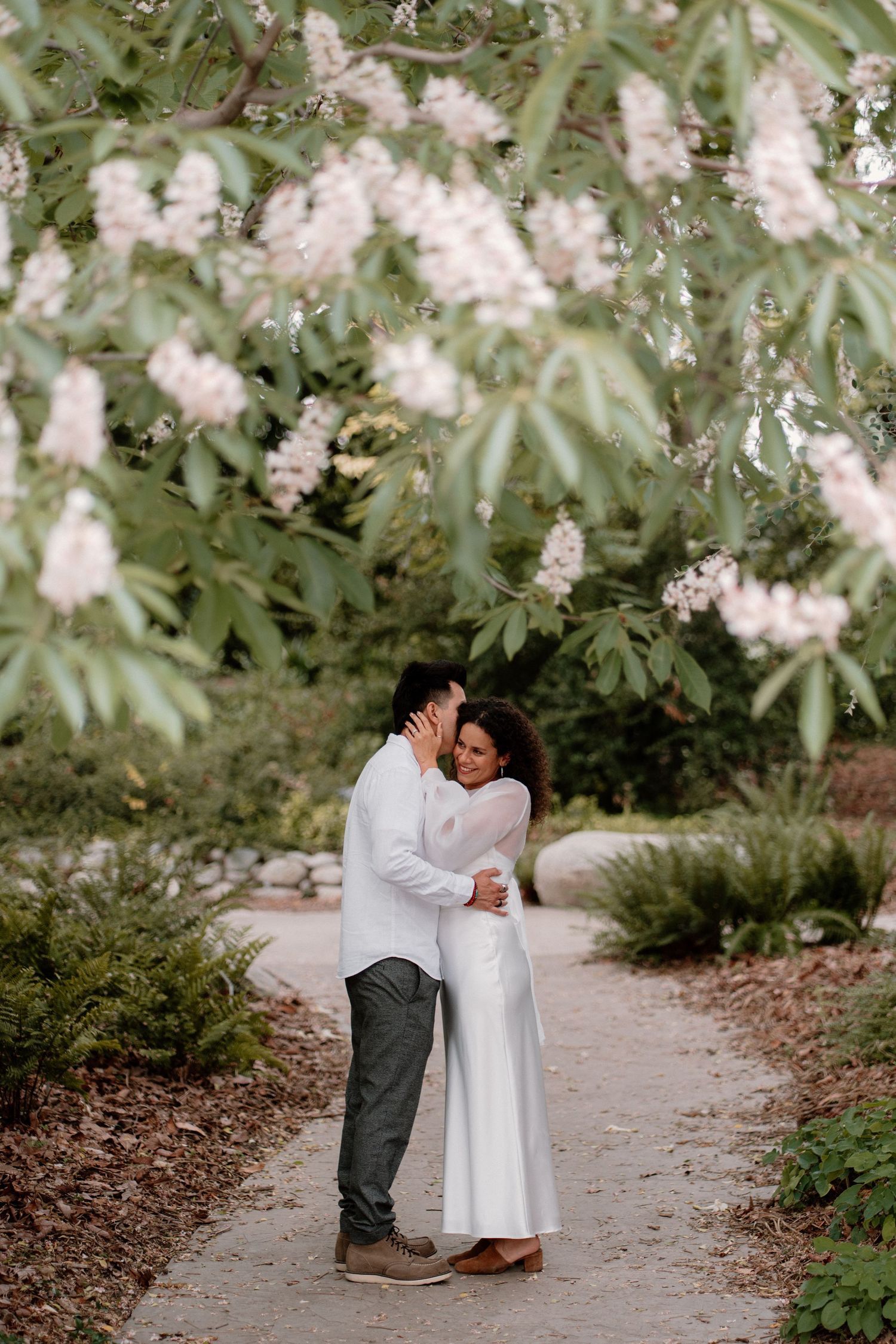 Couple embracing on a garden path under blooming white flowers during an engagement shoot.