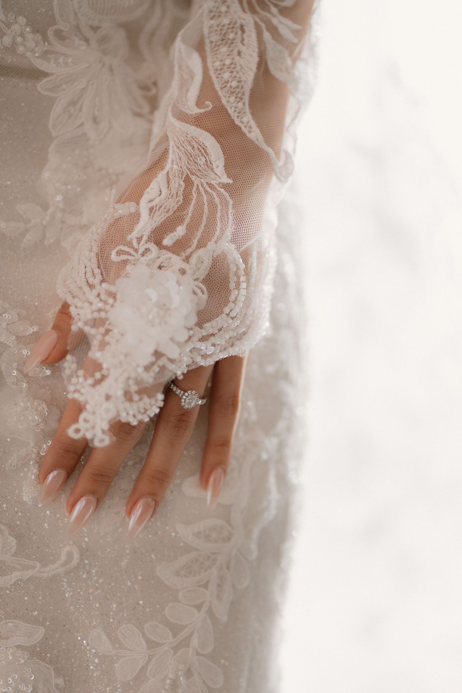 Close-up of a bride's hand adorned with an engagement ring wearing an intricate beaded lace wedding gown sleeve.