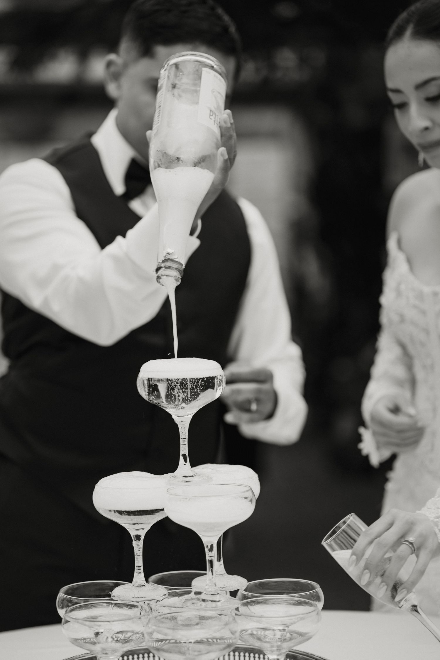 Groom pouring champagne tower at wedding reception in classic black and white photography style.