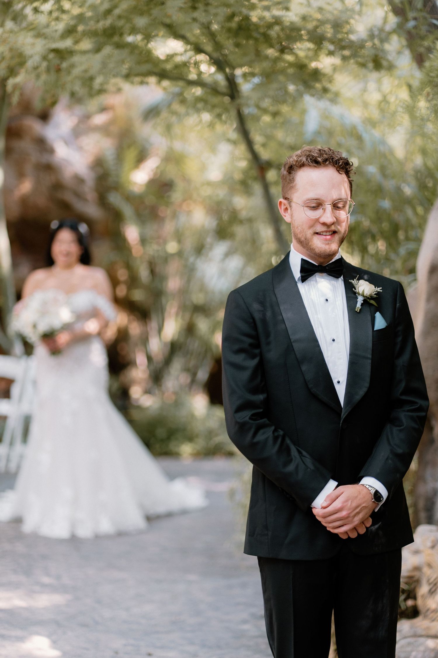 Emotional groom in black tuxedo awaits his bride during a first look moment in a lush garden setting.