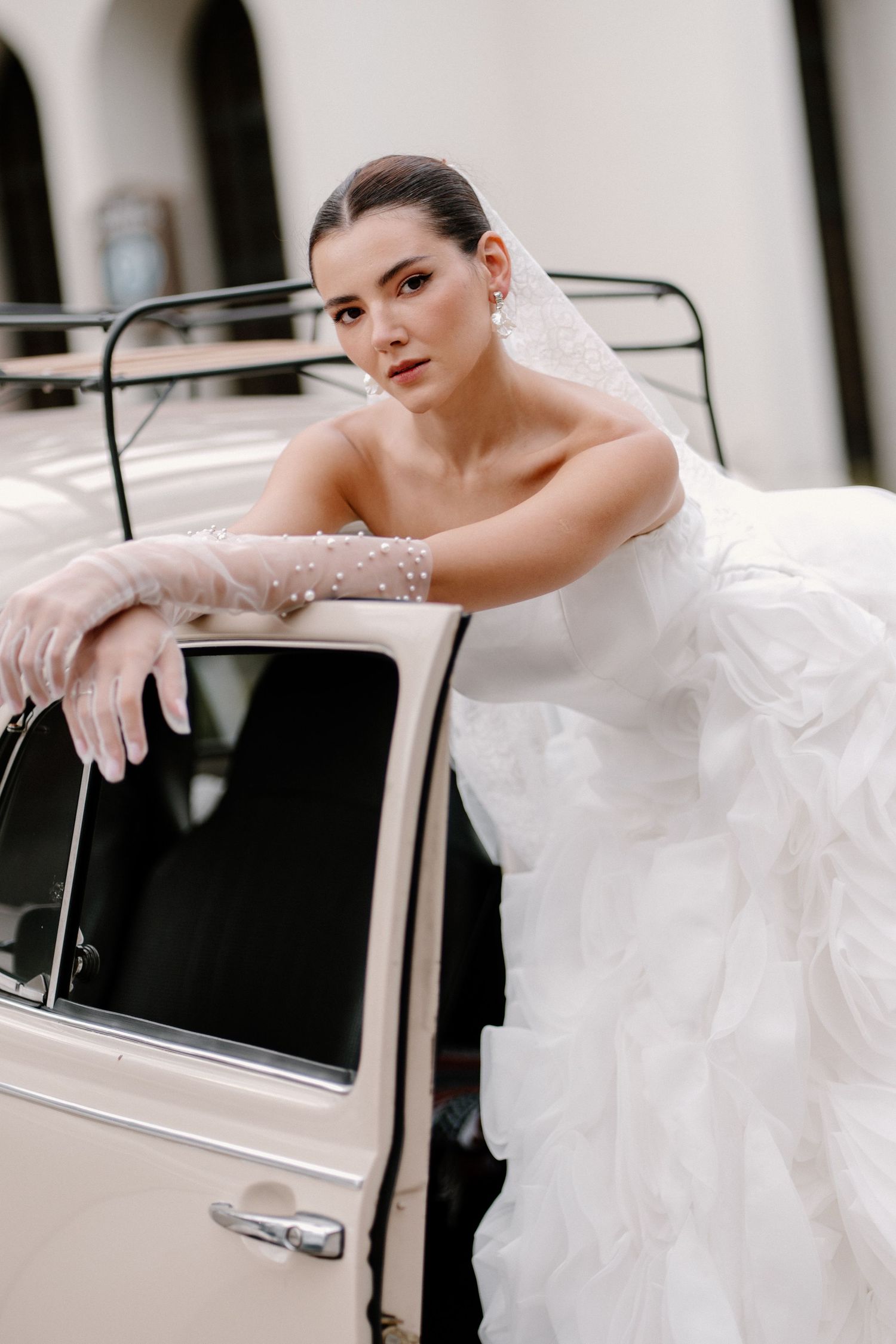 Elegant bride in strapless ruffled gown and pearl gloves leaning against vintage car.