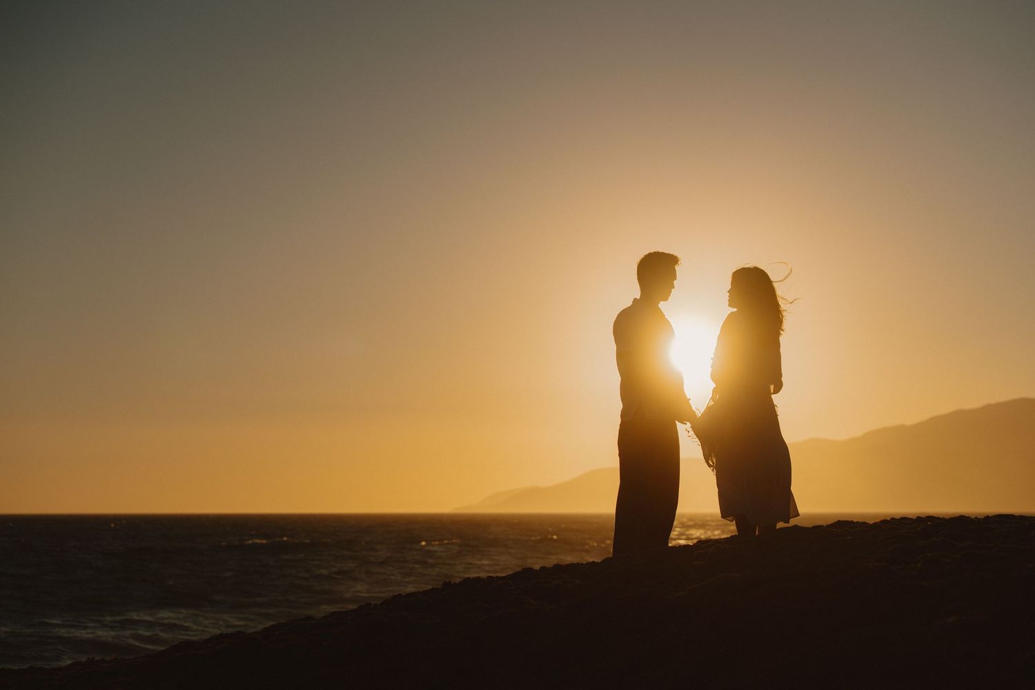 Romantic couple silhouetted against golden sunset on beach by ocean.