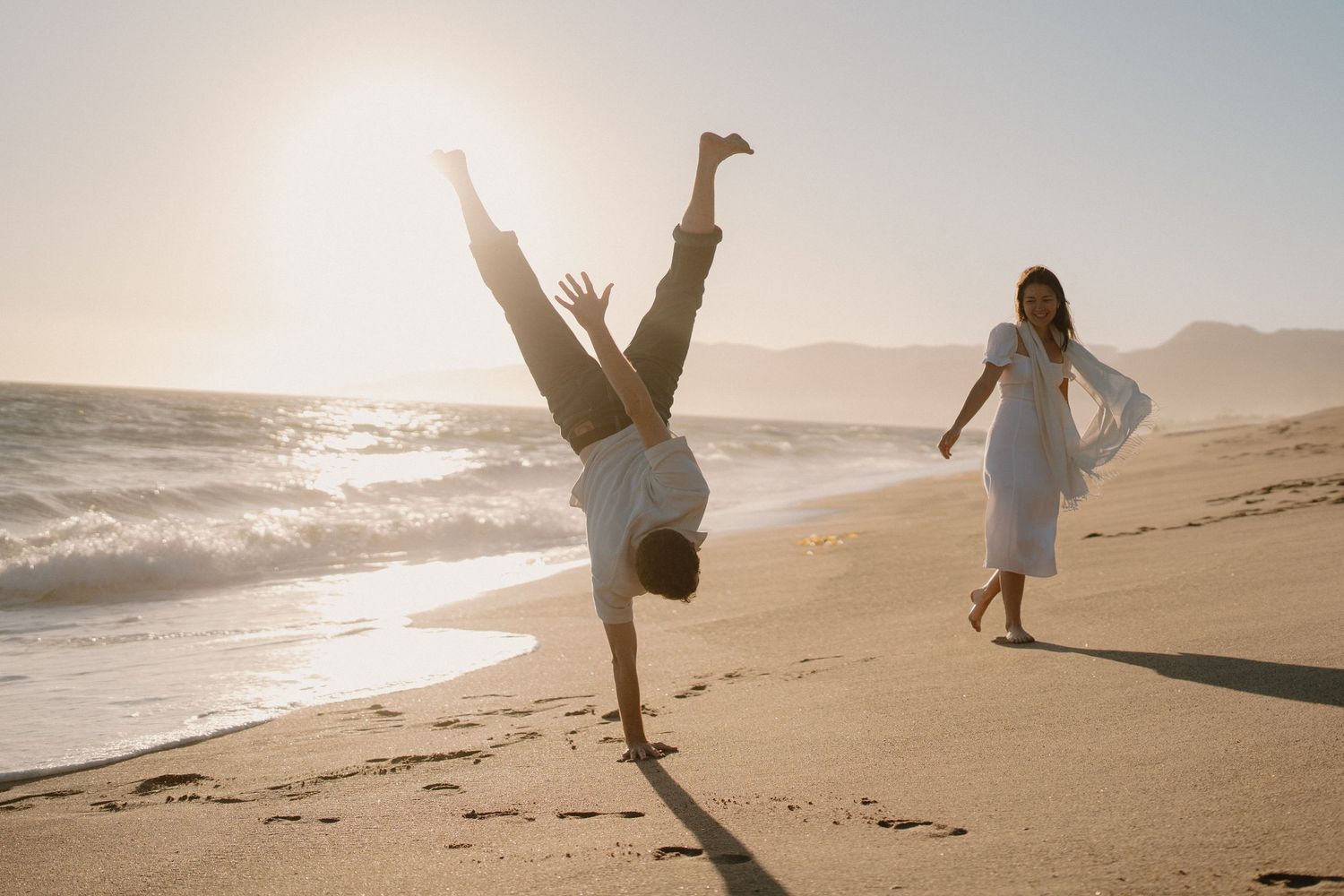 Man doing a cartwheel on a sunny beach while woman in white dress walks and laughs behind him.