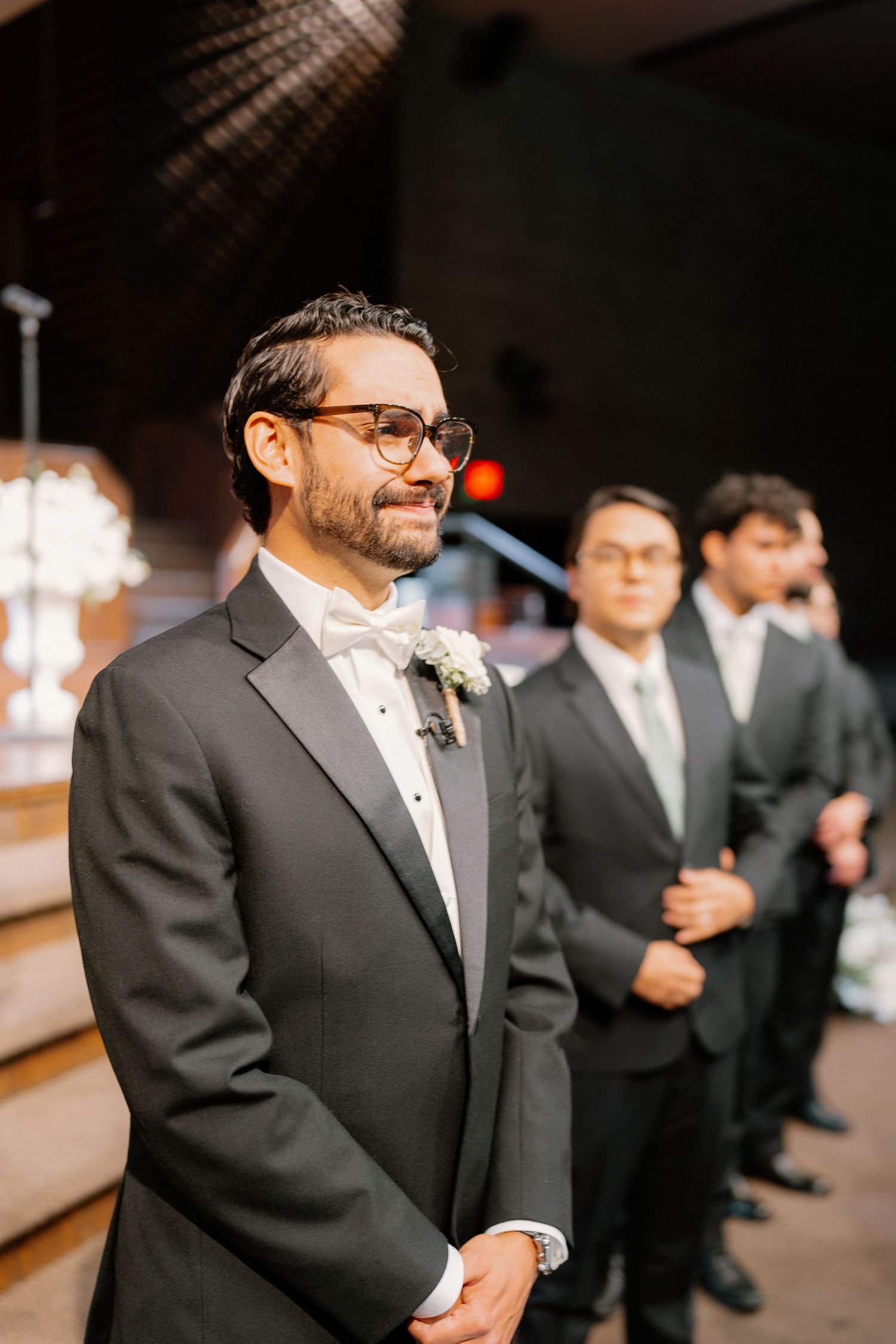 Smiling groom in black tuxedo with white bow tie and boutonniere stands at altar with groomsmen.