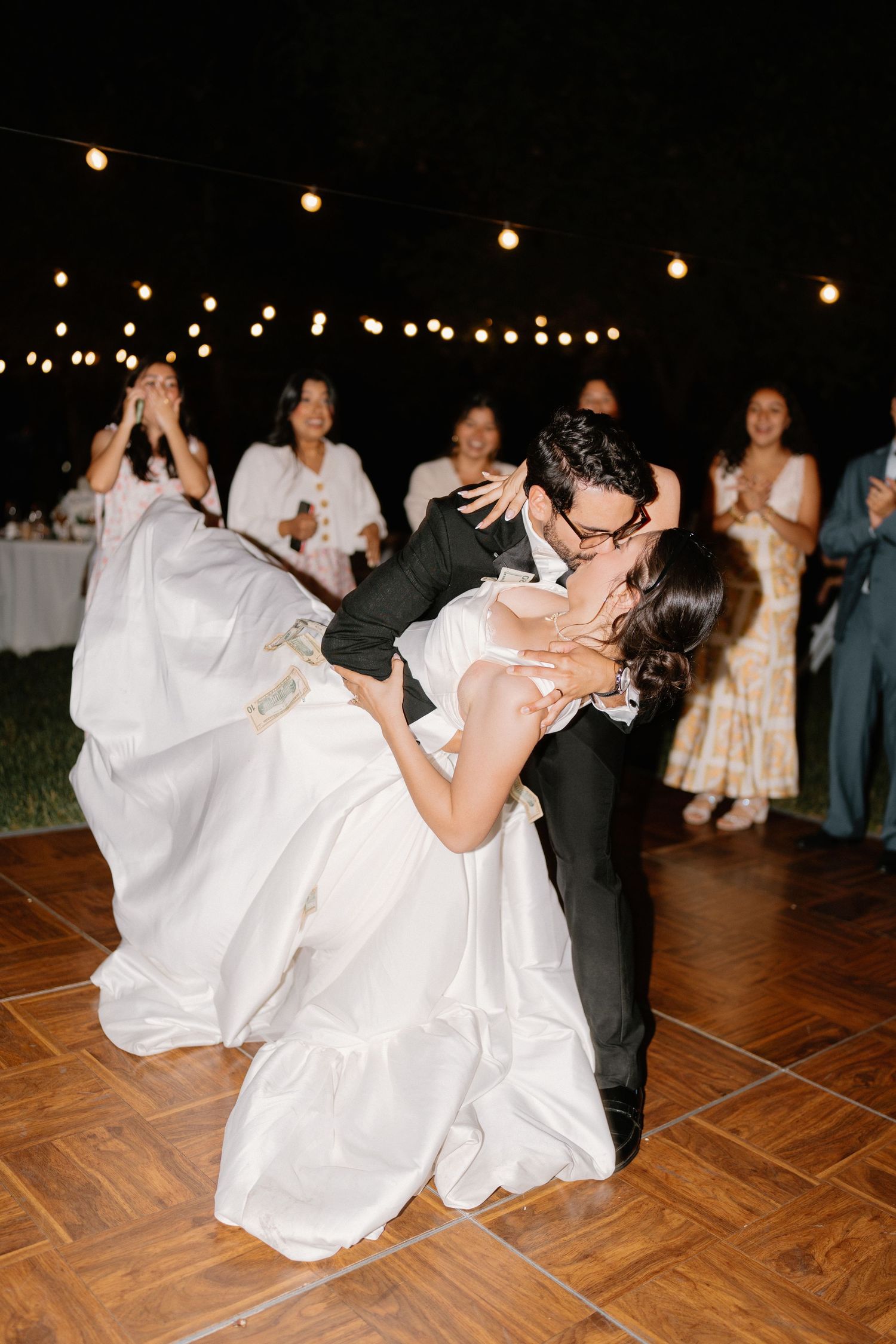 Groom dips bride in white gown for romantic kiss on outdoor dance floor under string lights.