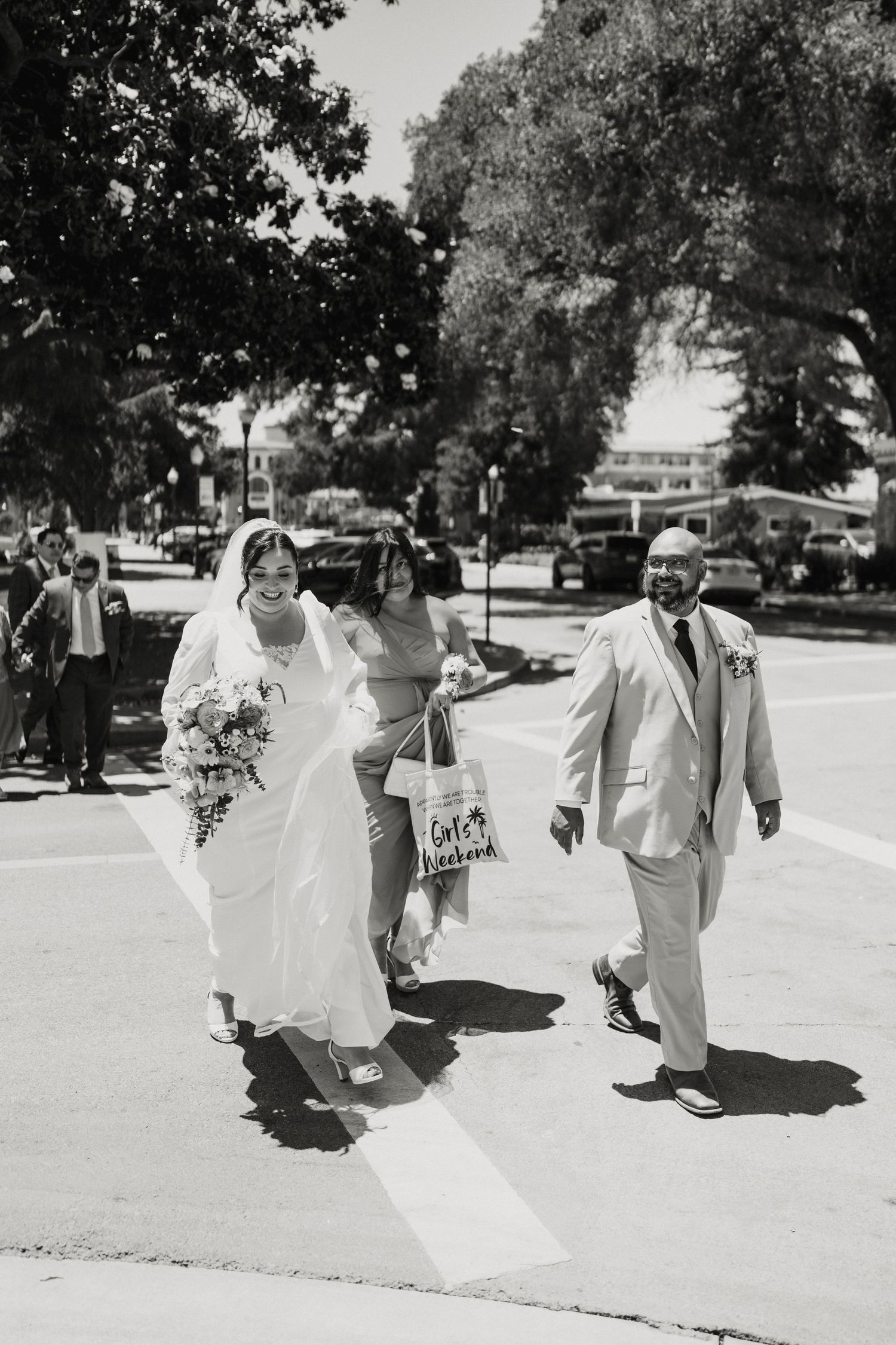 Bride in white gown and veil walks with groom in tan suit across sunny street on wedding day.
