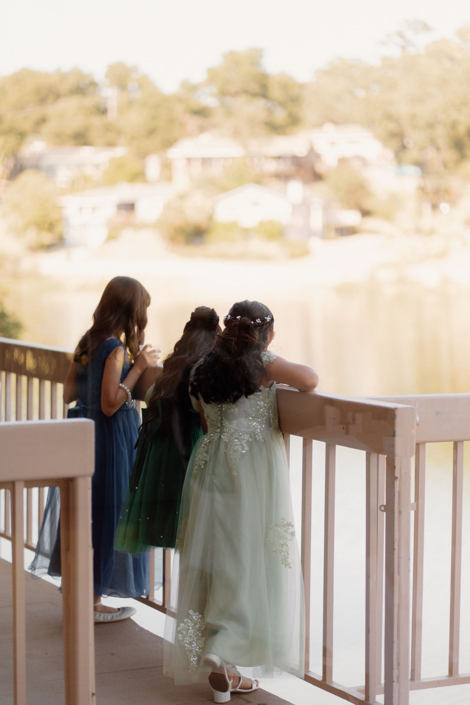 Three young girls in formal dresses standing at a railing overlooking a scenic waterway at a wedding.