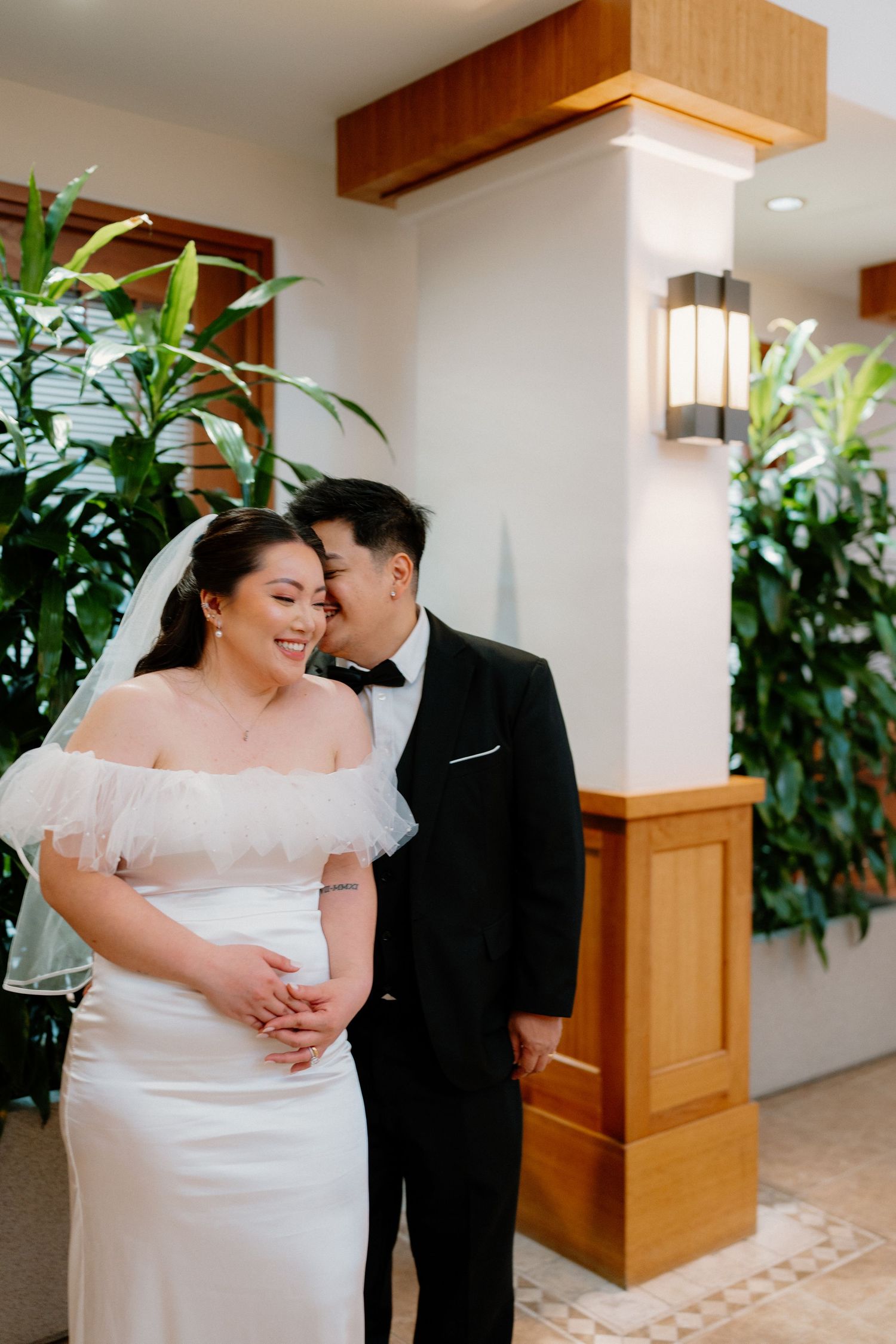 Groom whispers to smiling bride in white off-shoulder gown near lush indoor greenery.