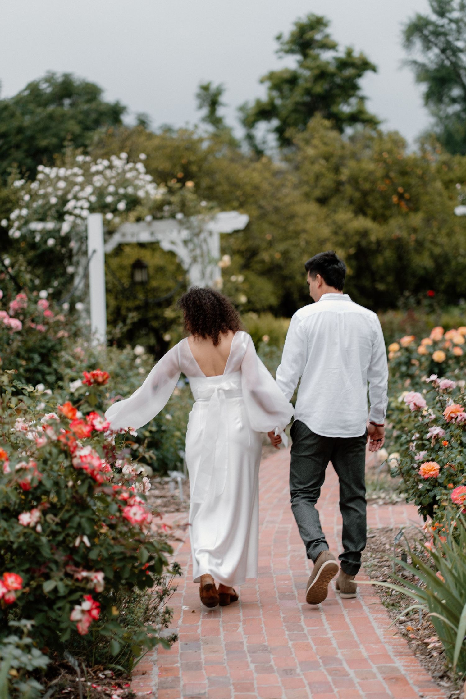 Couple walking hand in hand through a rose garden pathway, bride in white open-back dress.