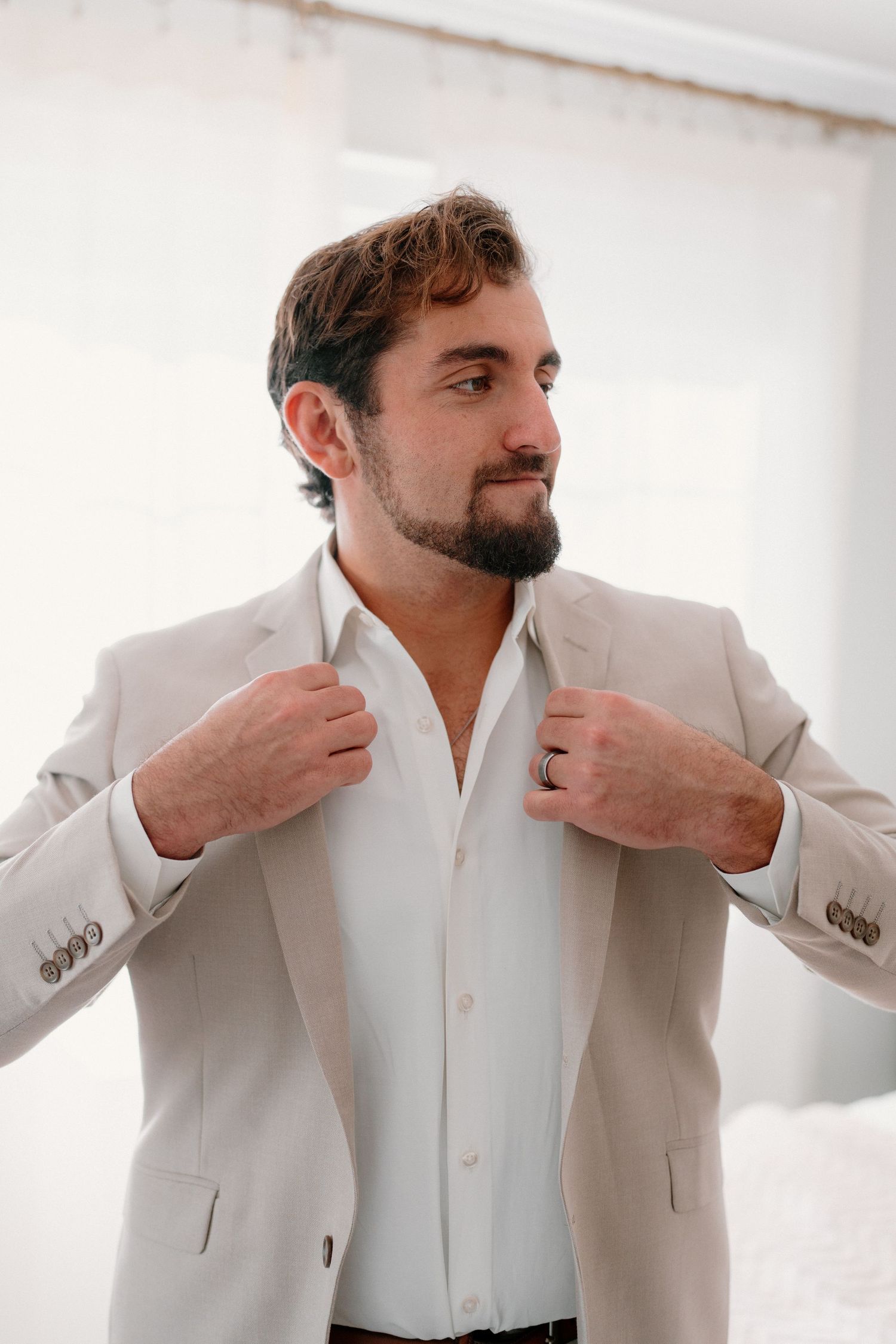 Handsome bearded groom in a beige suit adjusting his lapels while looking away in a bright white room.