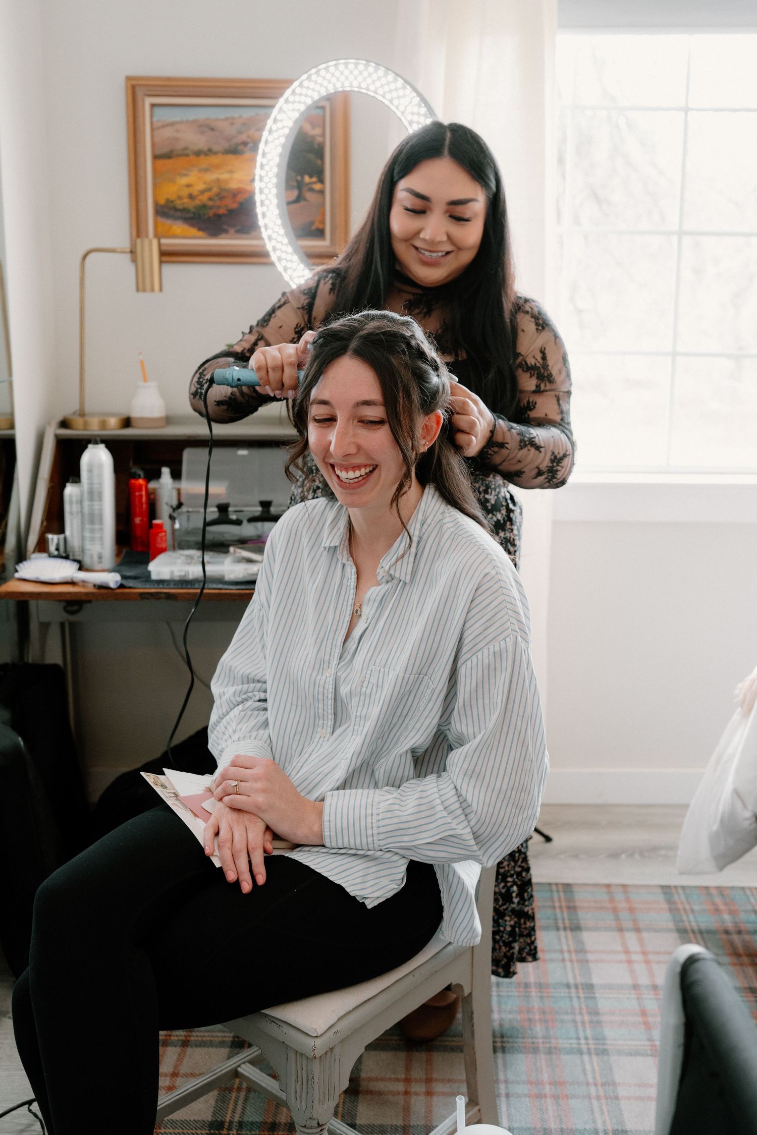 Smiling bride getting hair styled by hairdresser in bright salon room with ring light mirror.