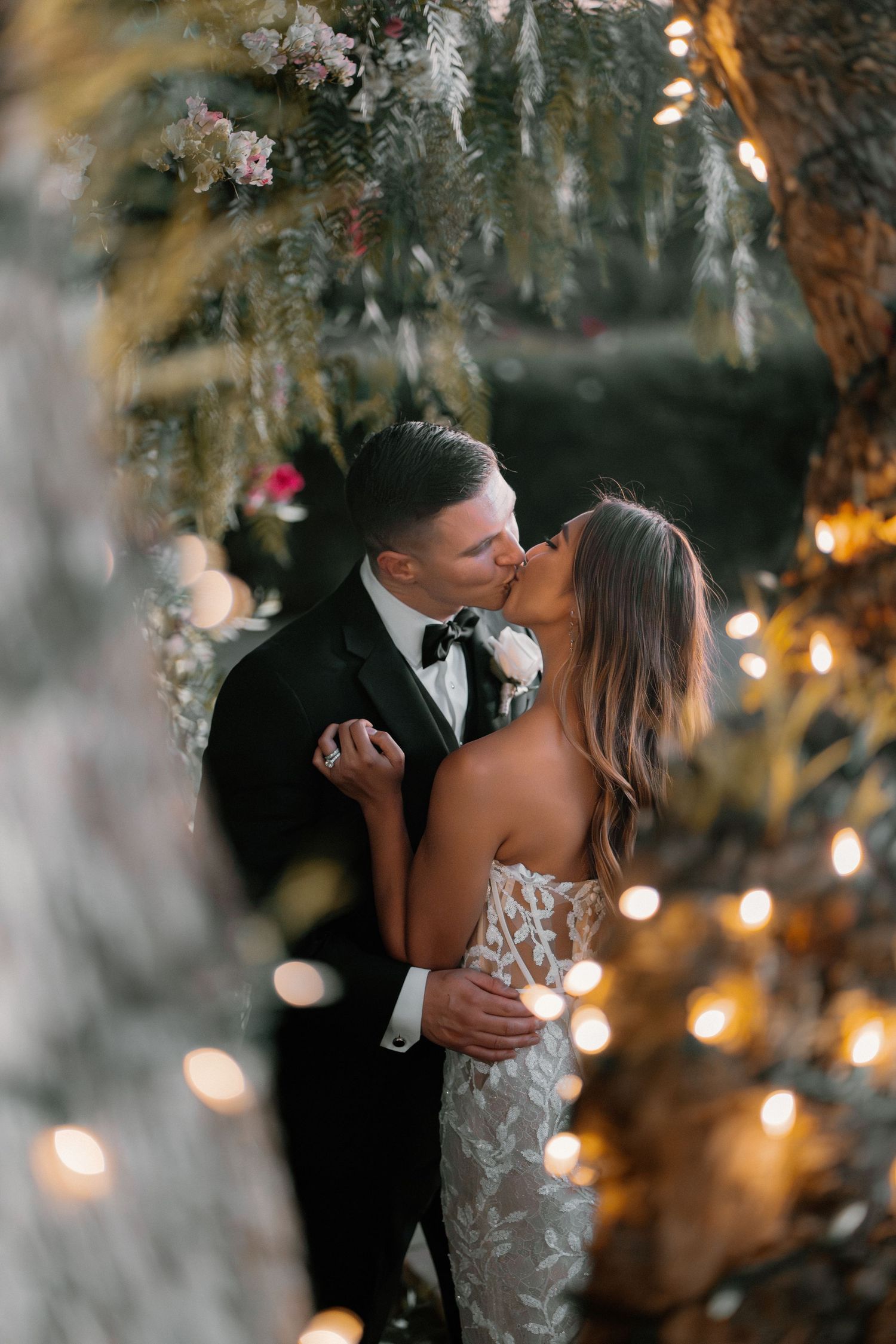 Bride and groom share romantic kiss surrounded by twinkling fairy lights and lush floral greenery.