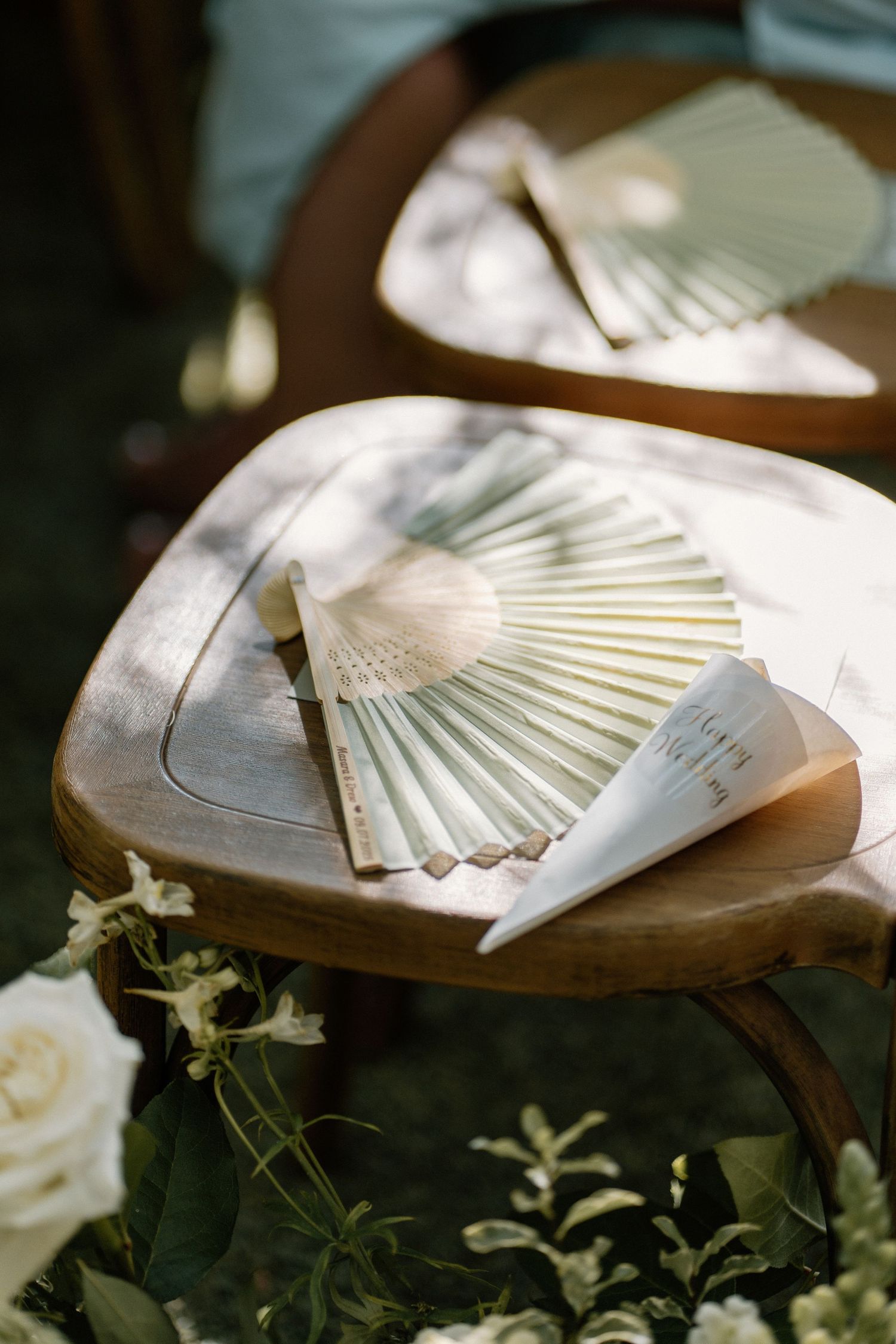 Elegant white paper hand fans with wedding programs on wooden chairs at outdoor ceremony.