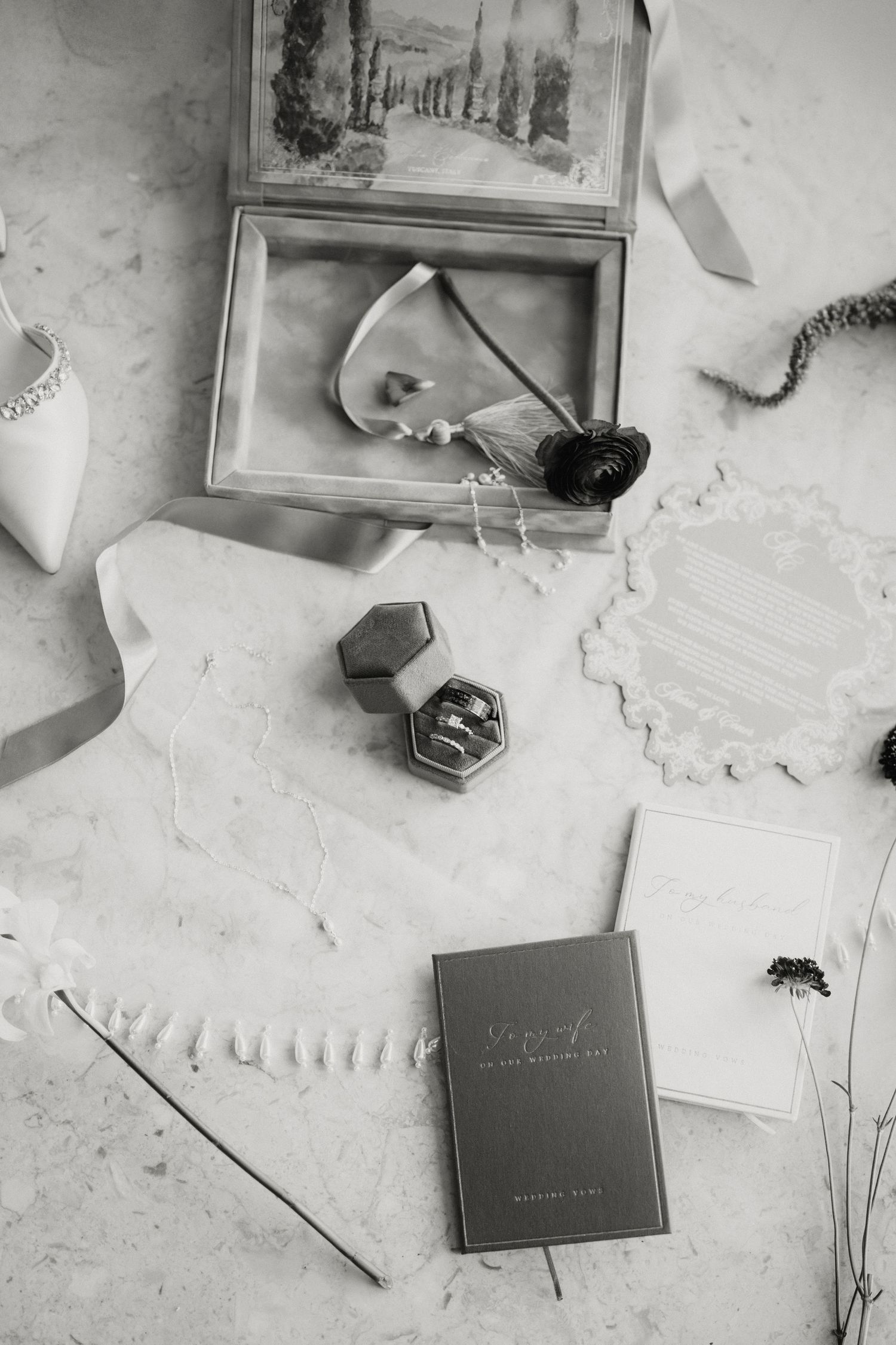 Black and white flat lay of wedding stationery, ring box, shoes, and floral accessories on marble.