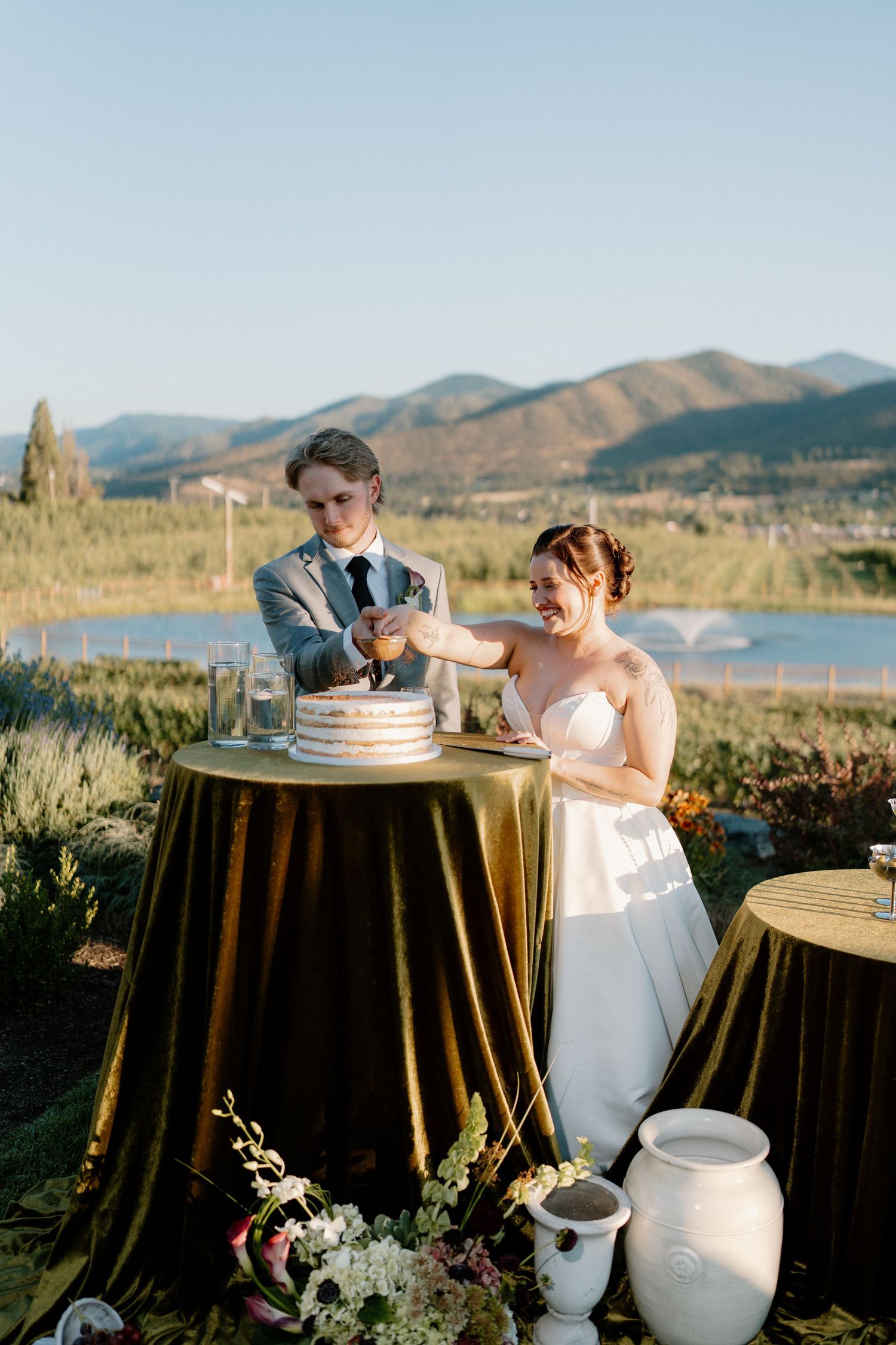 Bride and groom cut their naked wedding cake outdoors with mountain and lake scenery in background.